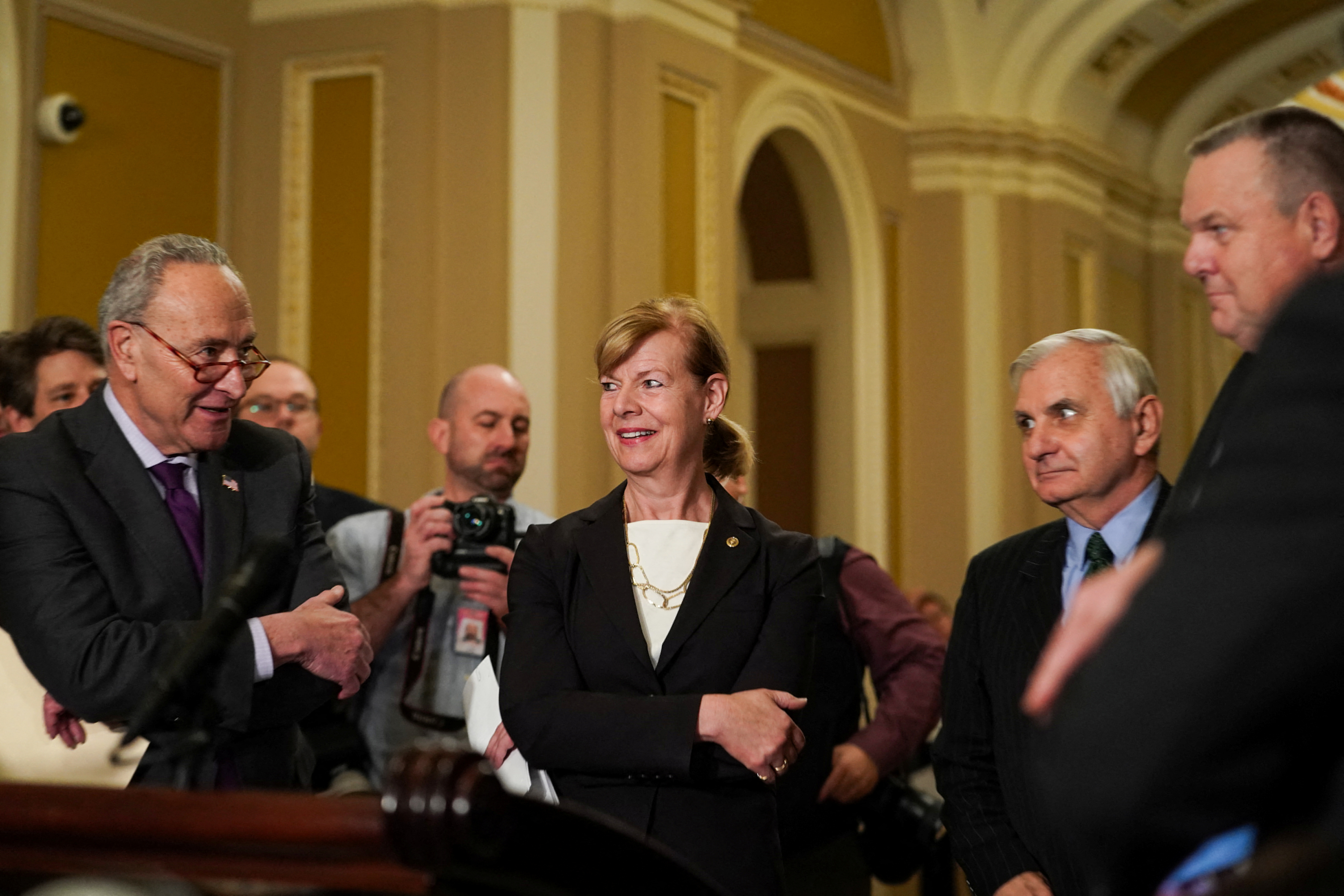 Chuck Schumer speaking to colleagues, all wearing black suit jackets, as a photographer stands behind them holding up camera