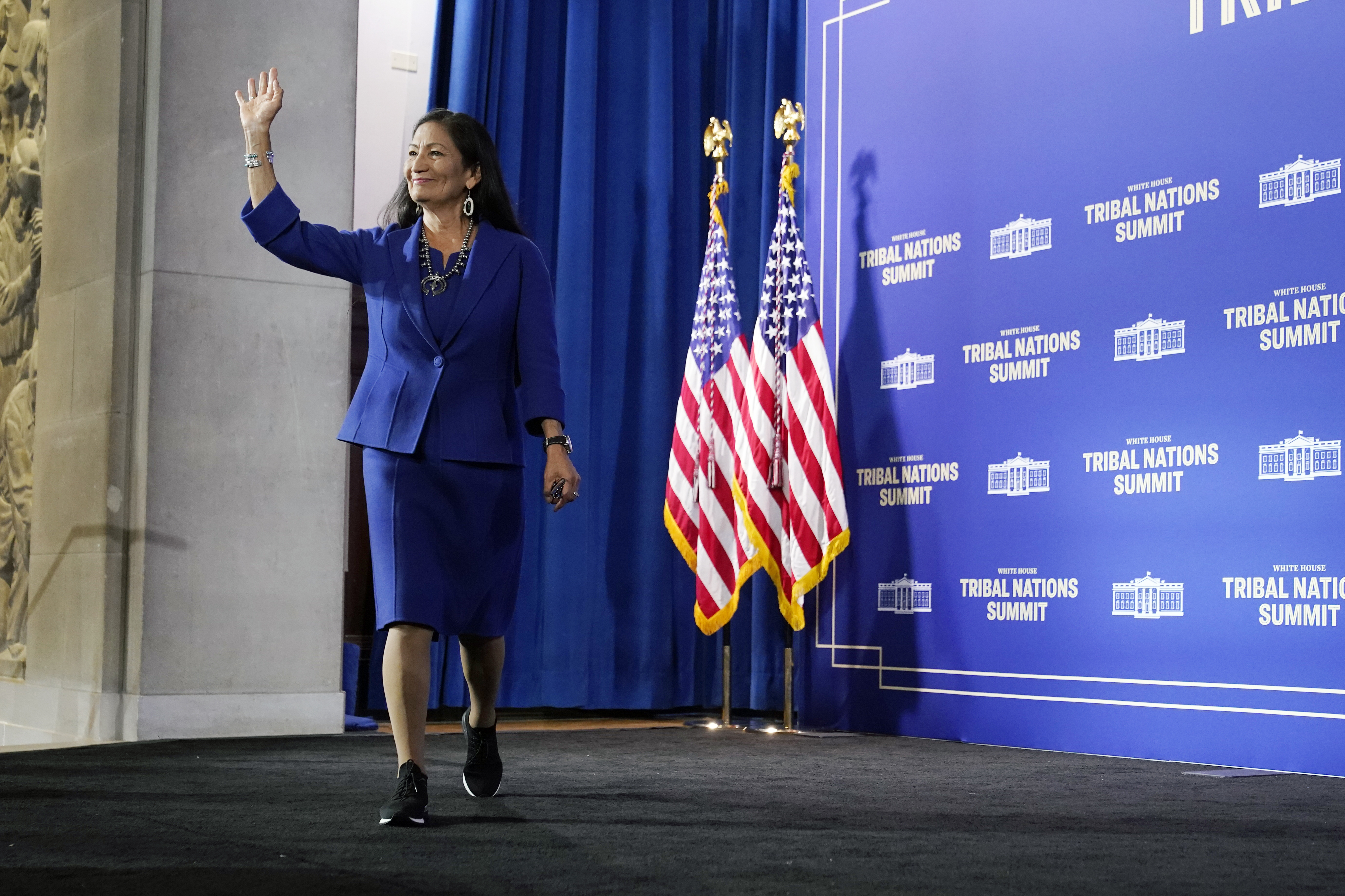 Deb Haaland waves from the stage as she walks forward, with two American flags behind her