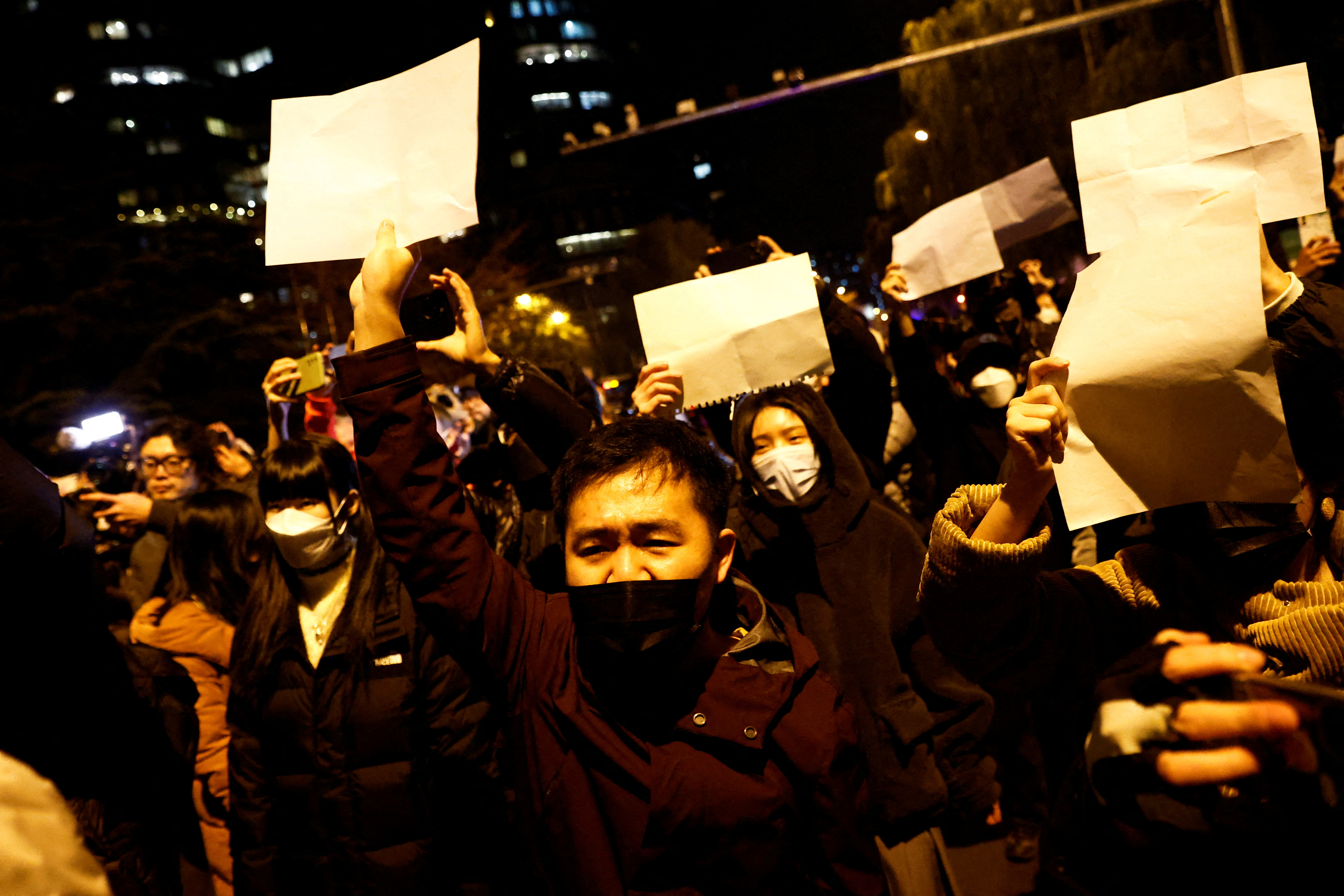 People demonstrate against coronavirus restrictions in the city of Urumqi. They are holding up pieces of paper with no writing on them. It's dark.