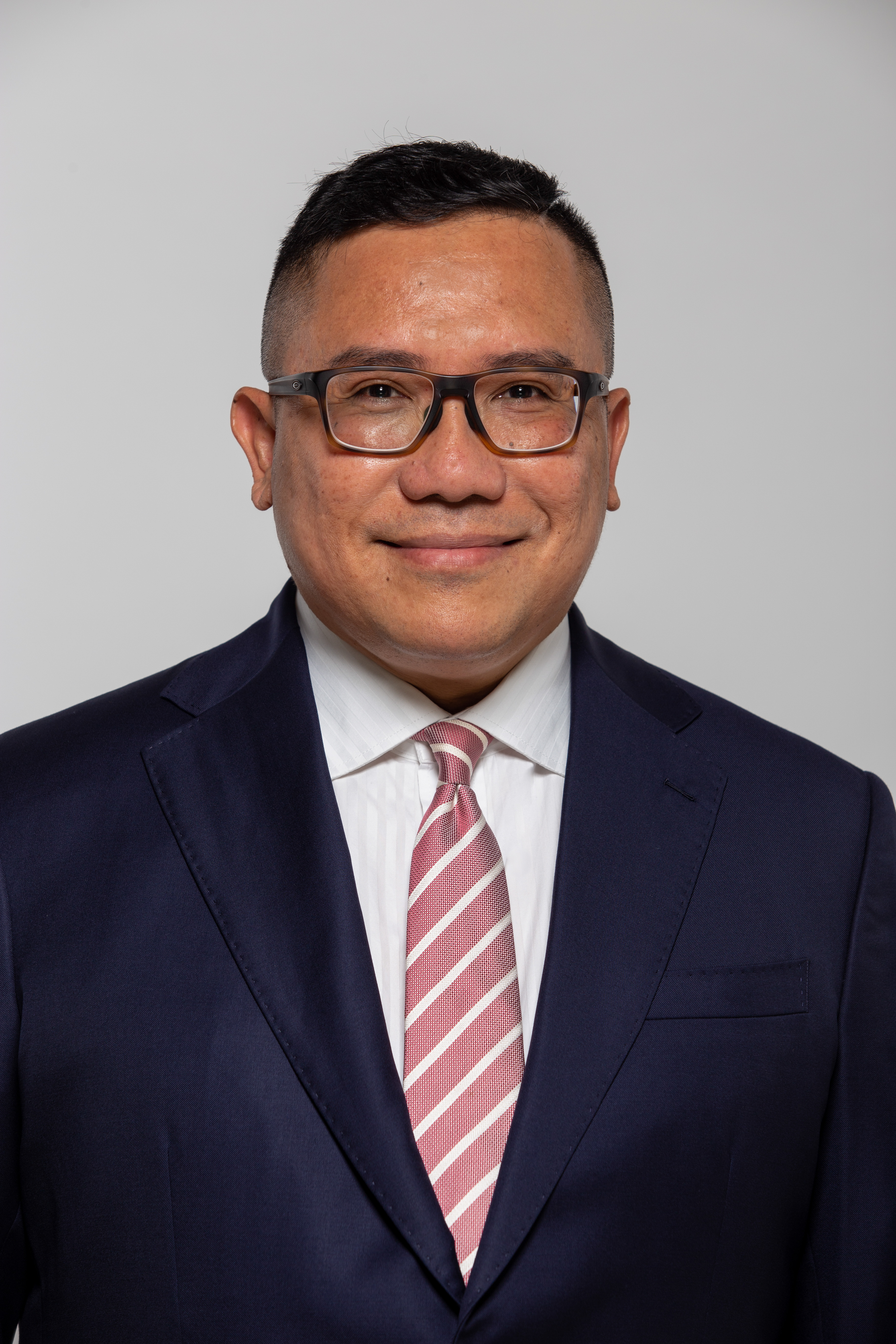 Ismail wearing glasses, with a short haircut, wearing a suit with a pink and white-striped tie