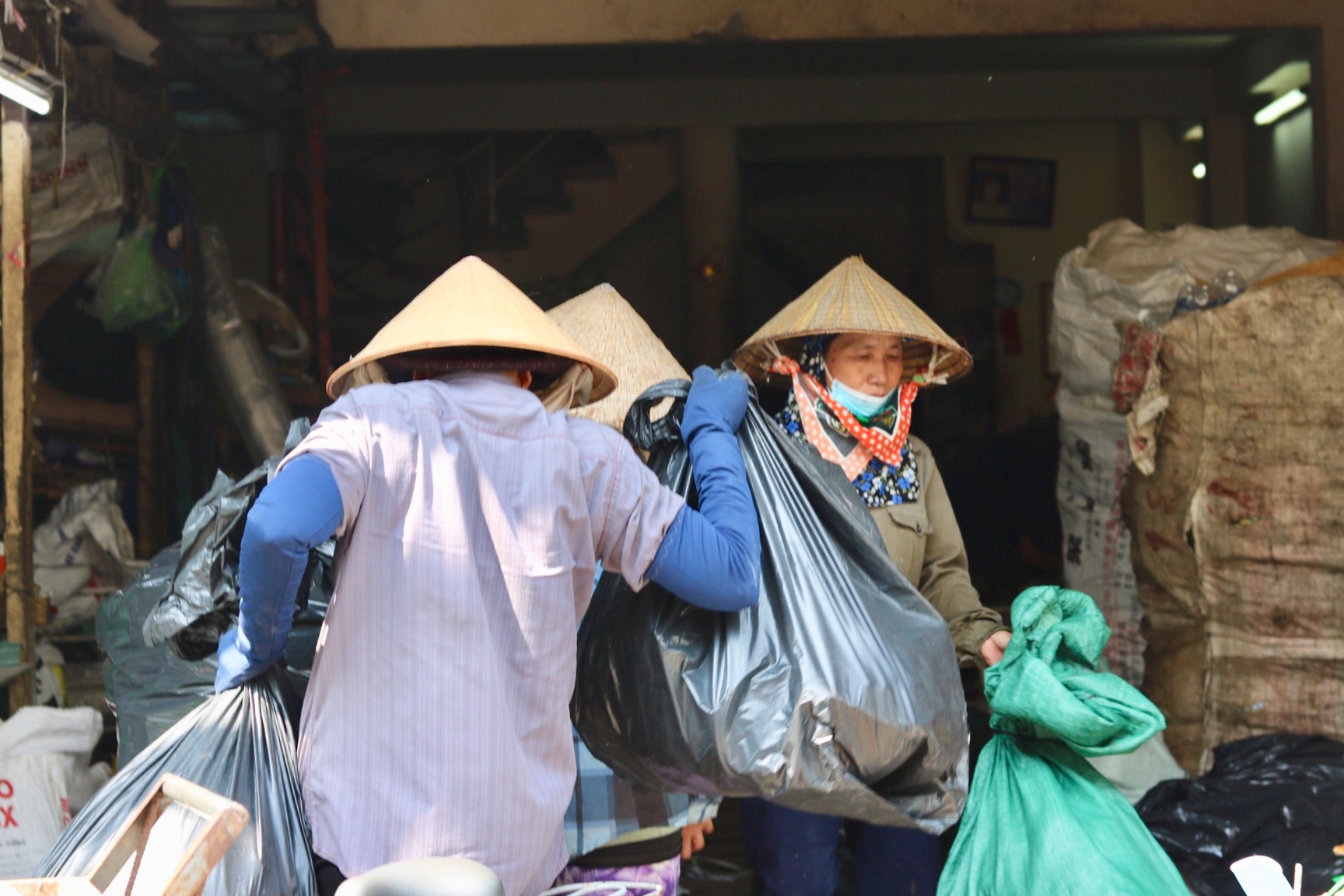 A waste collector in a conical hat walks into a collection centre with two large bags as a female collector walks out. She is also wearing a conical hat and is wearing a mask around her mouth. There are huge bags filled with waste to her left.