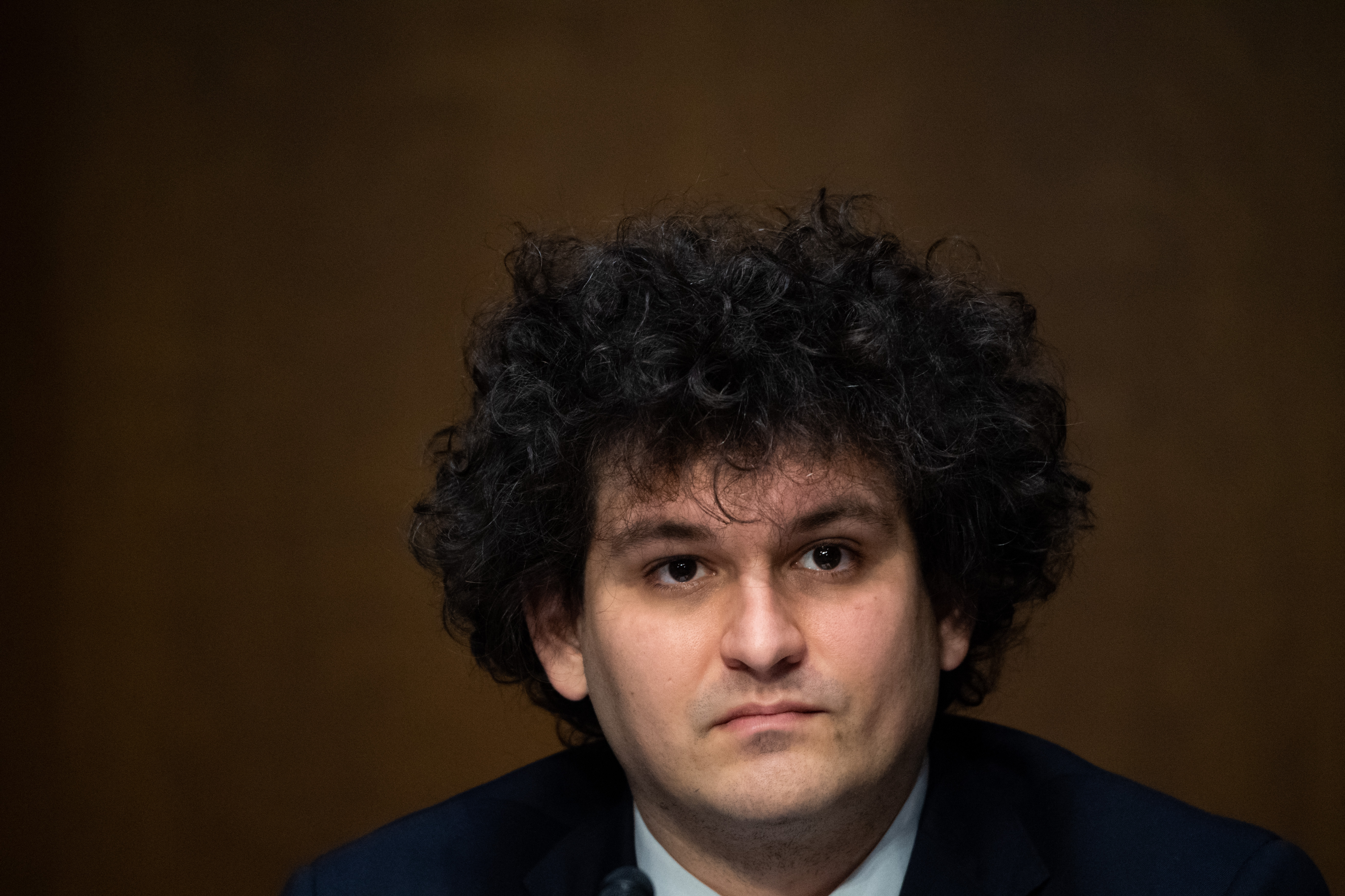 A portrait of Sam Bankman-Fried. He has dark, curly hair and looks serious