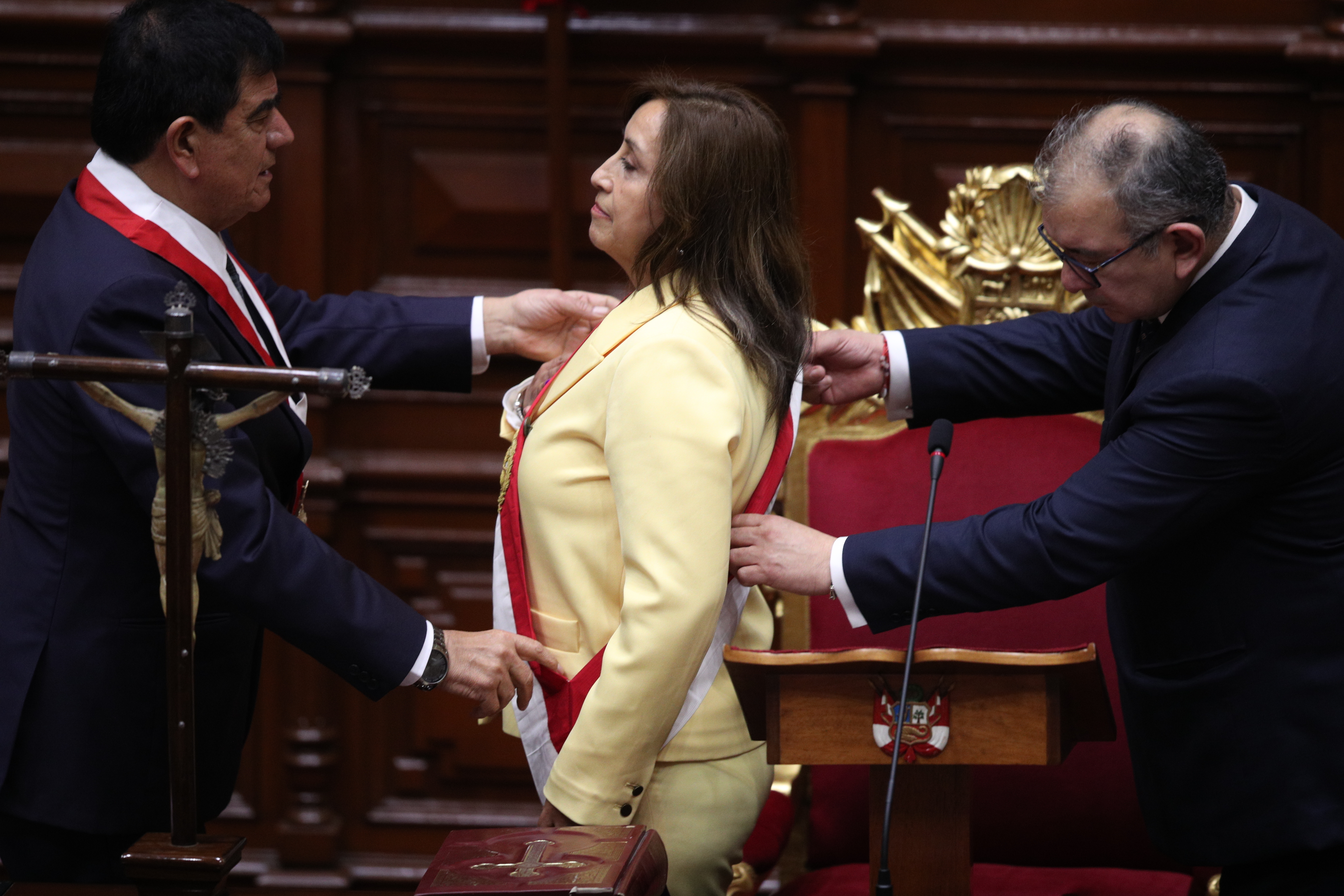 Boluarte stands in pale yellow suit two men, on either side, fix a red and white sash over her shoulder