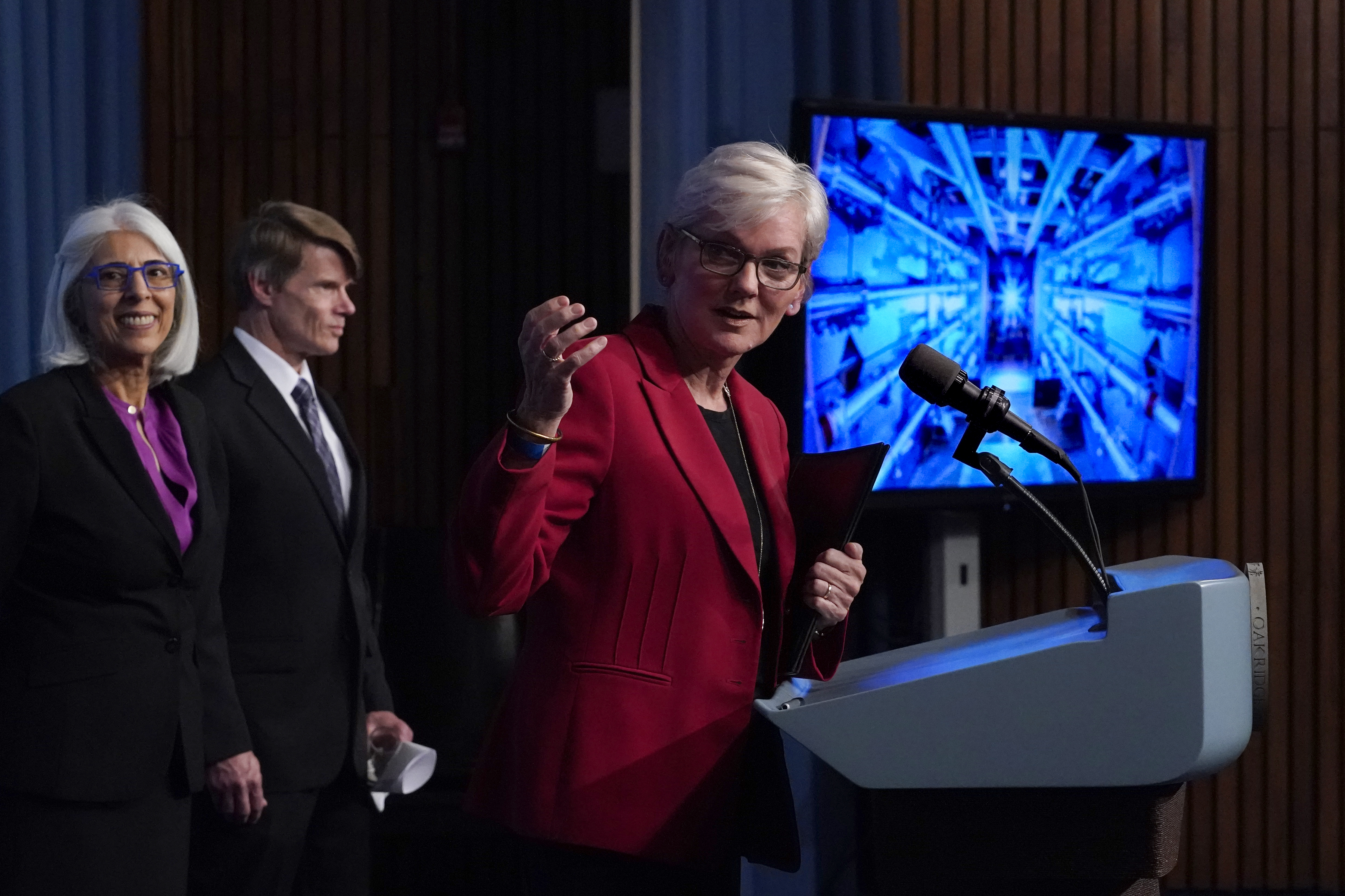 Secretary of Energy Jennifer Granholm, in a red suit announces the fusion breakthrough. There is a screen behind her.