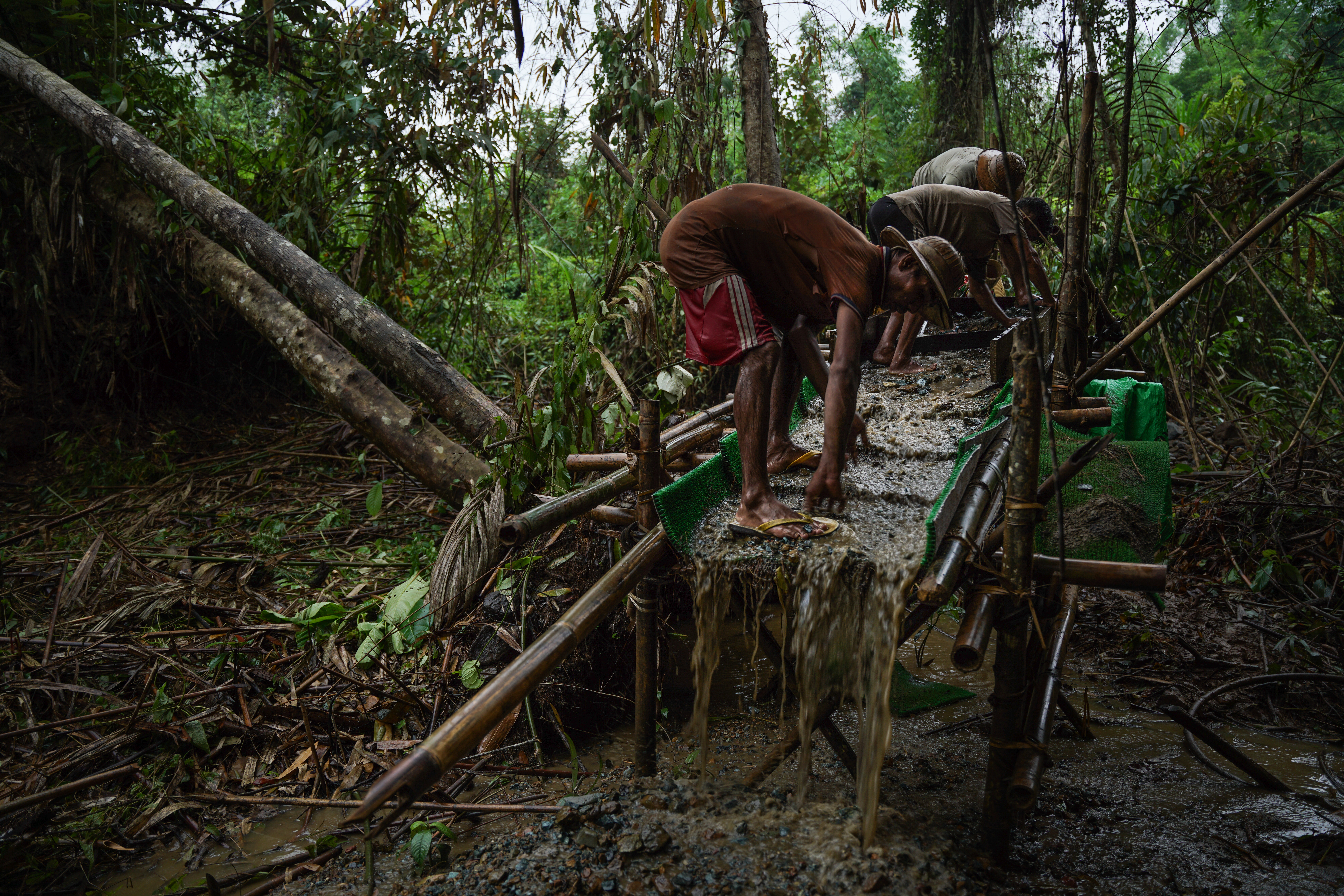 Three men standing on a contraption made of bamboo and netting that is filtering the river water for gold. There is jungle around them