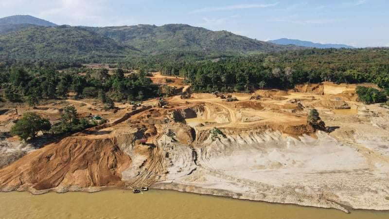 Gold mines in Myitsone seen from above. The land is cleared and left with only one or two trees. There are tracks through the mud down to the river. The river water, like the earth, is orangey brown. There are forests and hills in the background