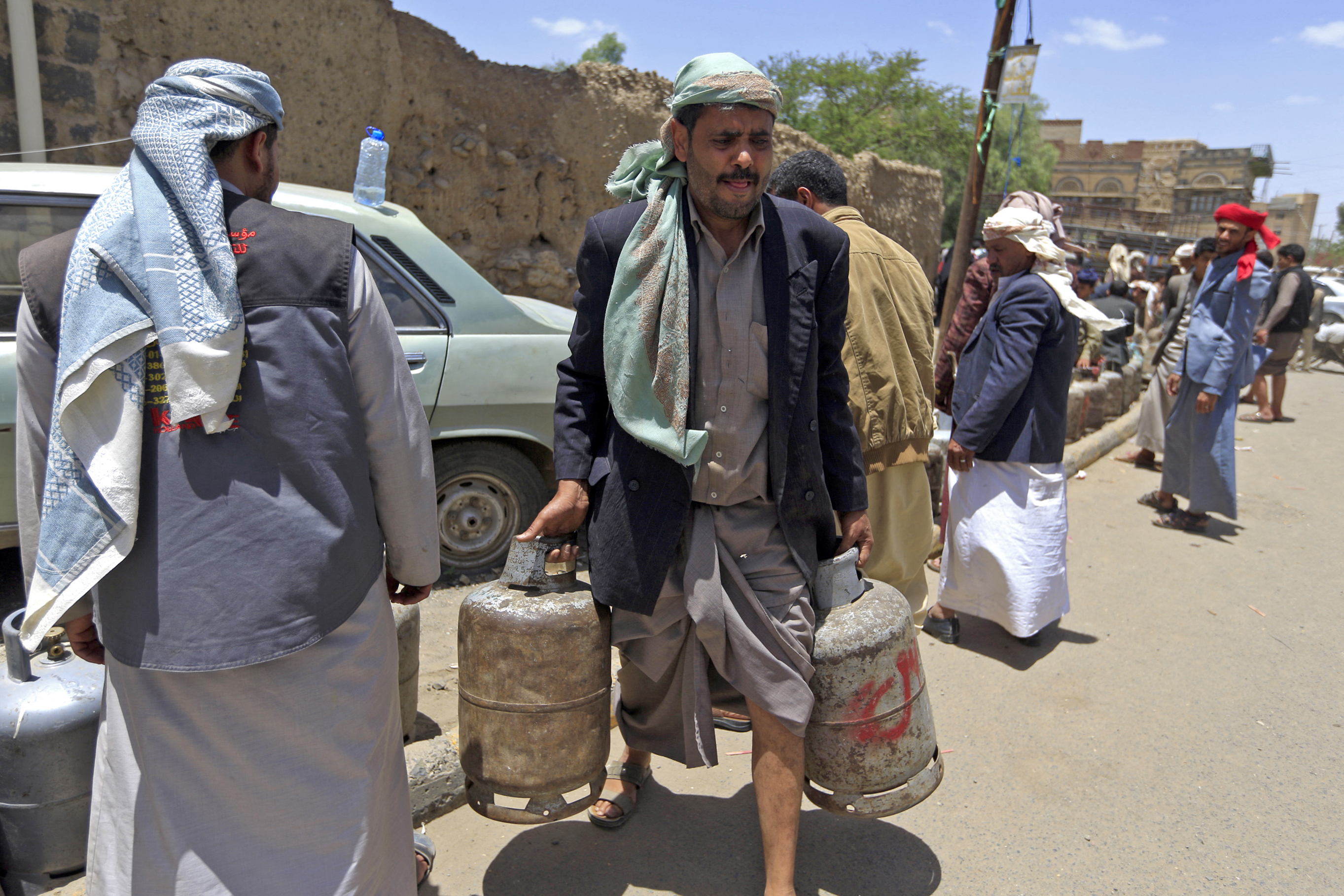 A photo of a man carrying two gas cylinders with people and a row of gas cylinders in the background.
