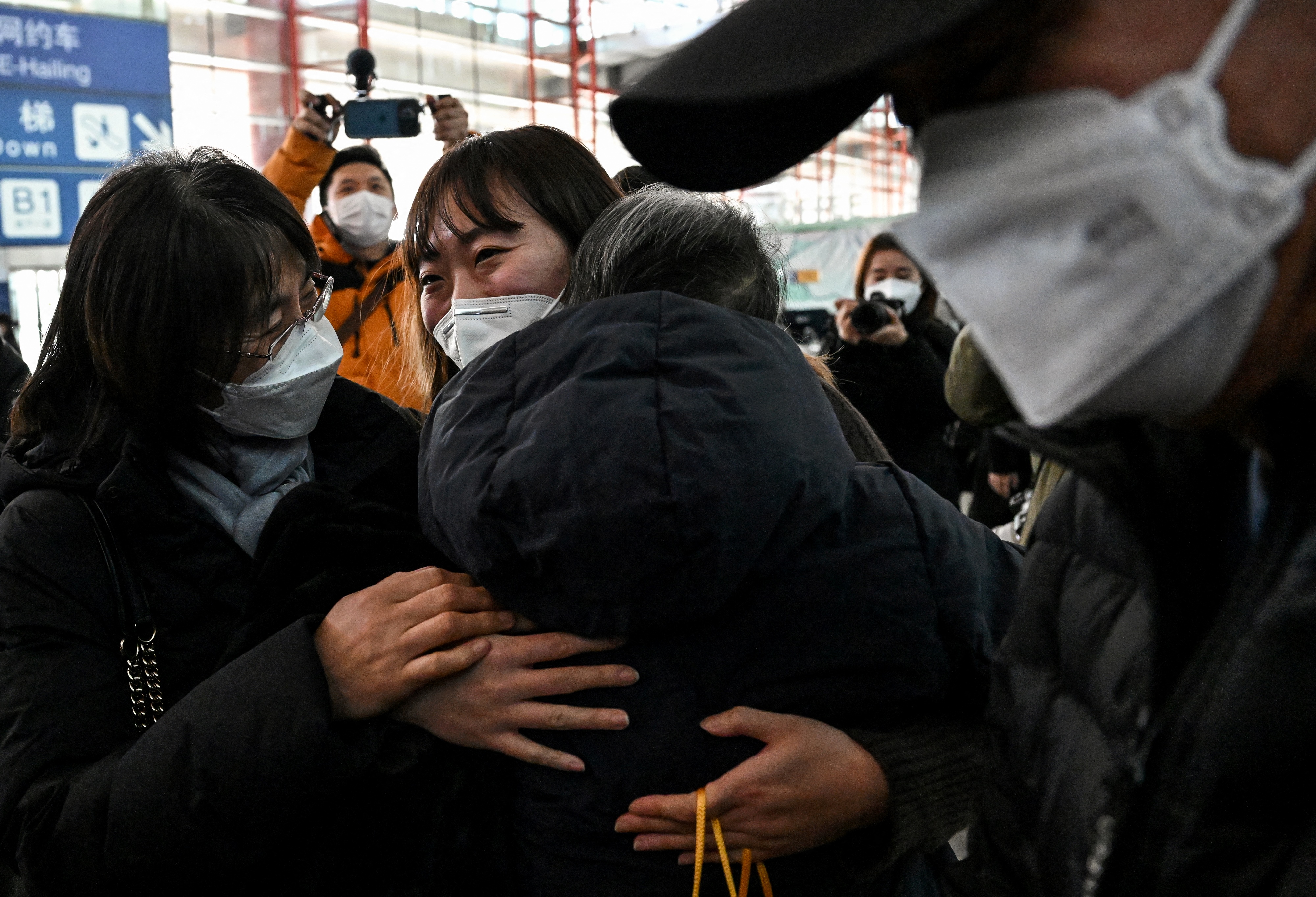 A woman in a face mask ehugs two other people at the international arrivals area of Beijing airport, She looks emotional. A man in a face mask is walking into the picture from the right. Everyone is wearing thick winter coats
