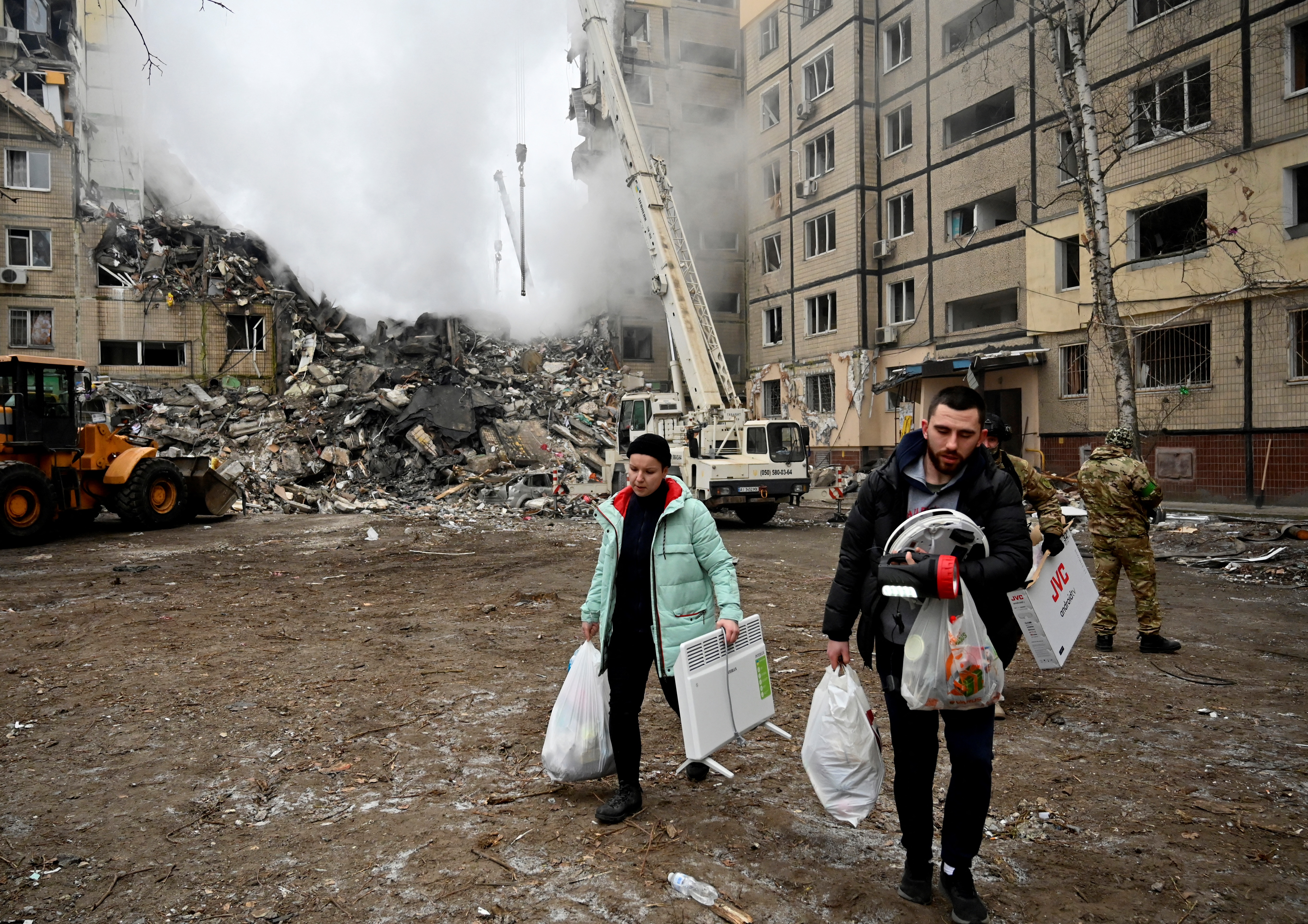 Two residents walk away from the smouldering ruins of a partially destroyed apartment building, with white plastic bags of belongings in their hands.