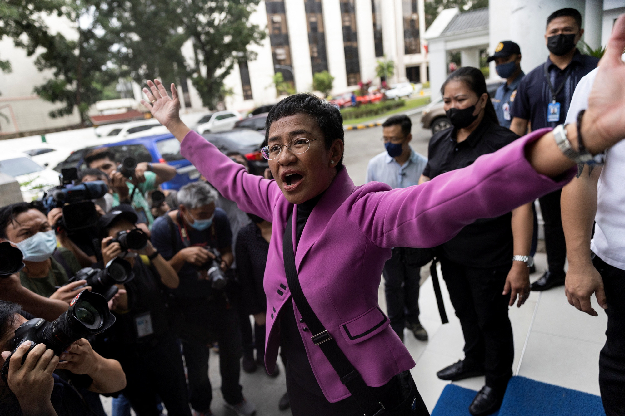 Maria Ressa wearing a purple jacket has her arms out wide outside the court, looking jubilant
