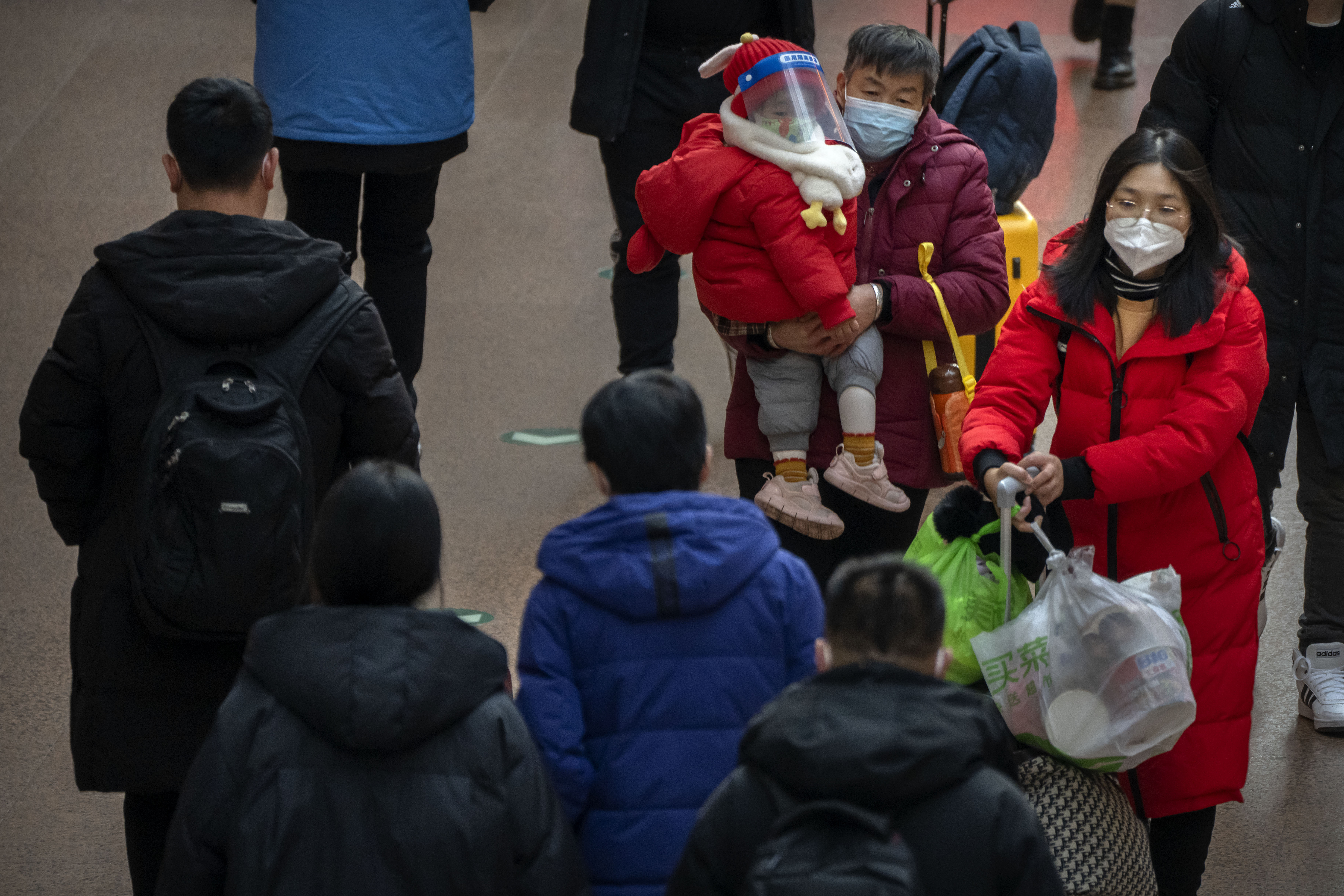 A woman carries a child wearing a face mask and face shield as they walk along a concourse at Beijing West Railway Station in Beijing