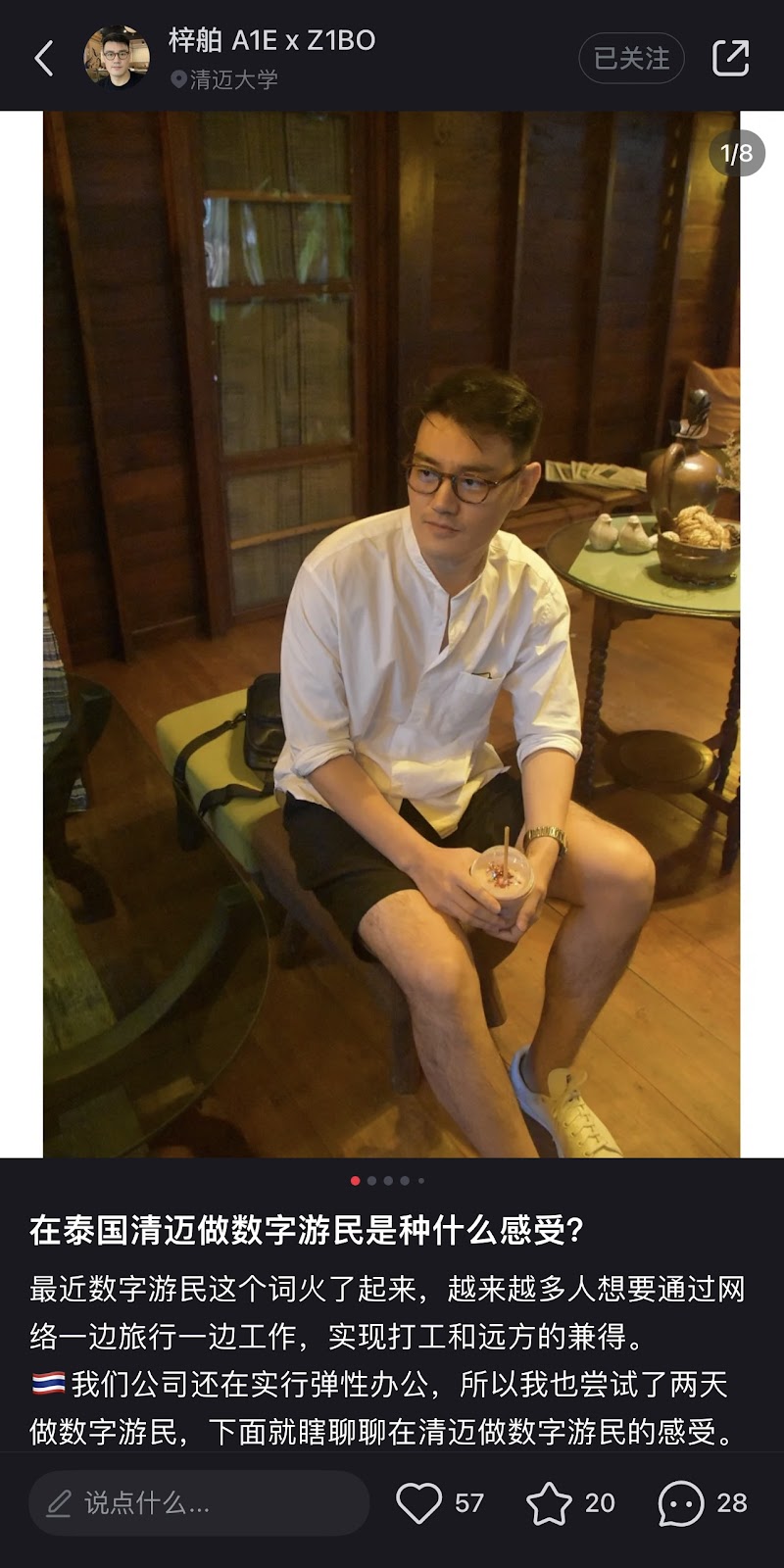 Man wearing a white short, rolled up to the elbows, and black shorts sits on n ottoman holding a drink with a straw in his hands.