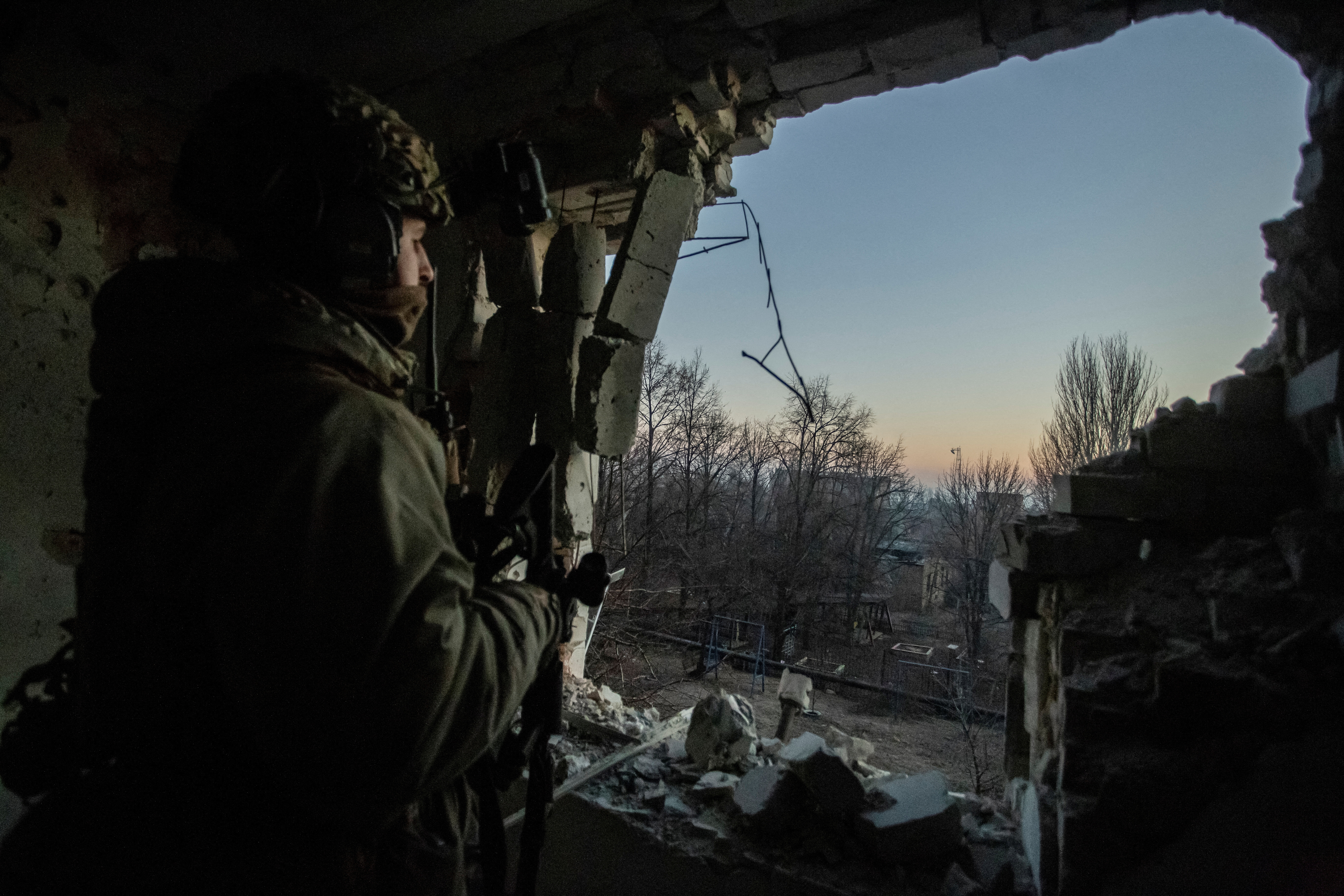 Ukrainian soldier stands in a cave, looking out towards the opening that shows a clear dimming blue sky, with a setting sun over naked trees on the horizon. It looks cold