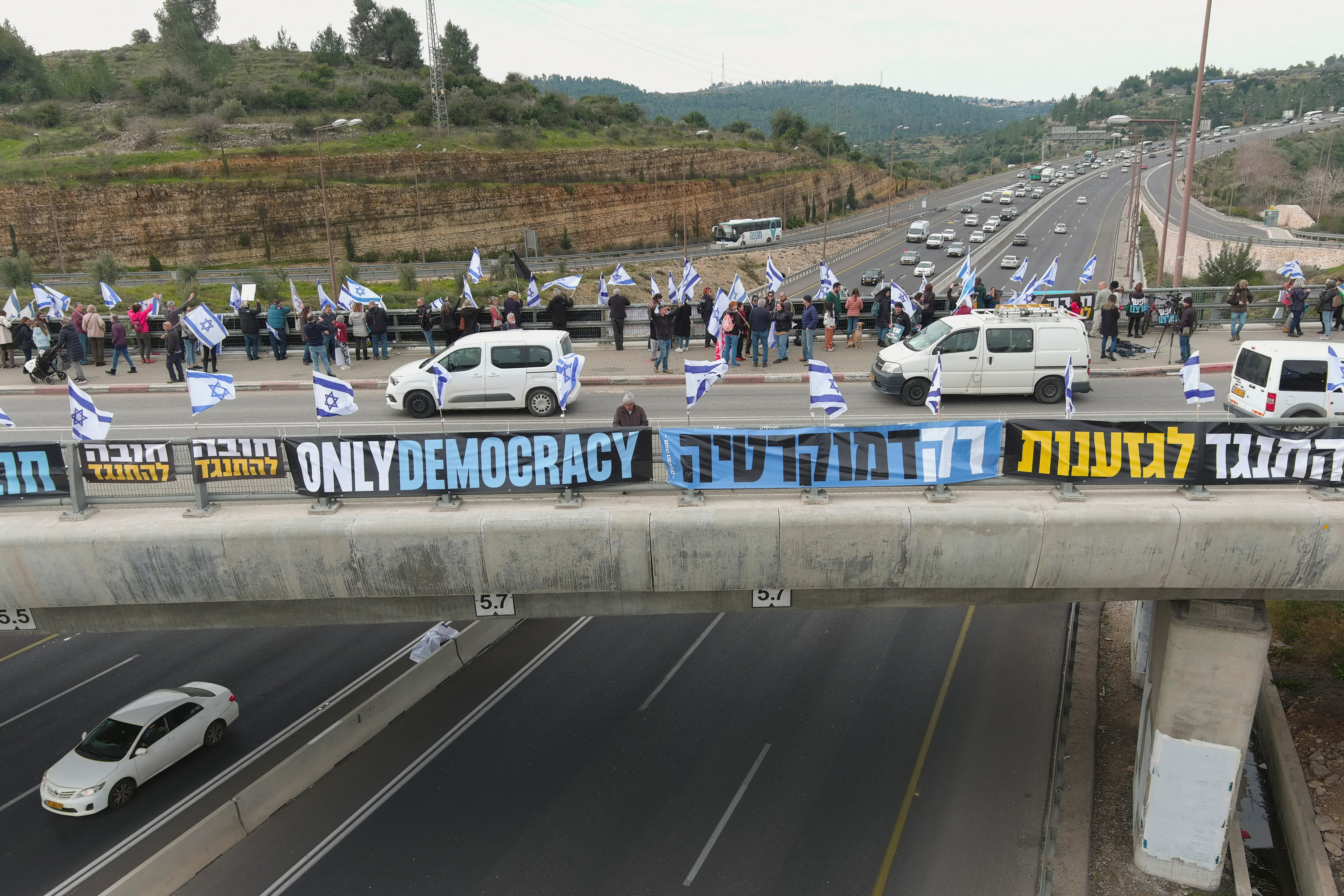 An aerial views shows Israelis holding flags as they demonstrate on a bridge on the day Israel's constitution committee is set to start voting on changes that would give politicians more power on selecting judges while limiting Supreme Court powers to strike down legislation, outside the Knesset, Israel's parliament, at Hemed Junction in Ein Hemed, February 13