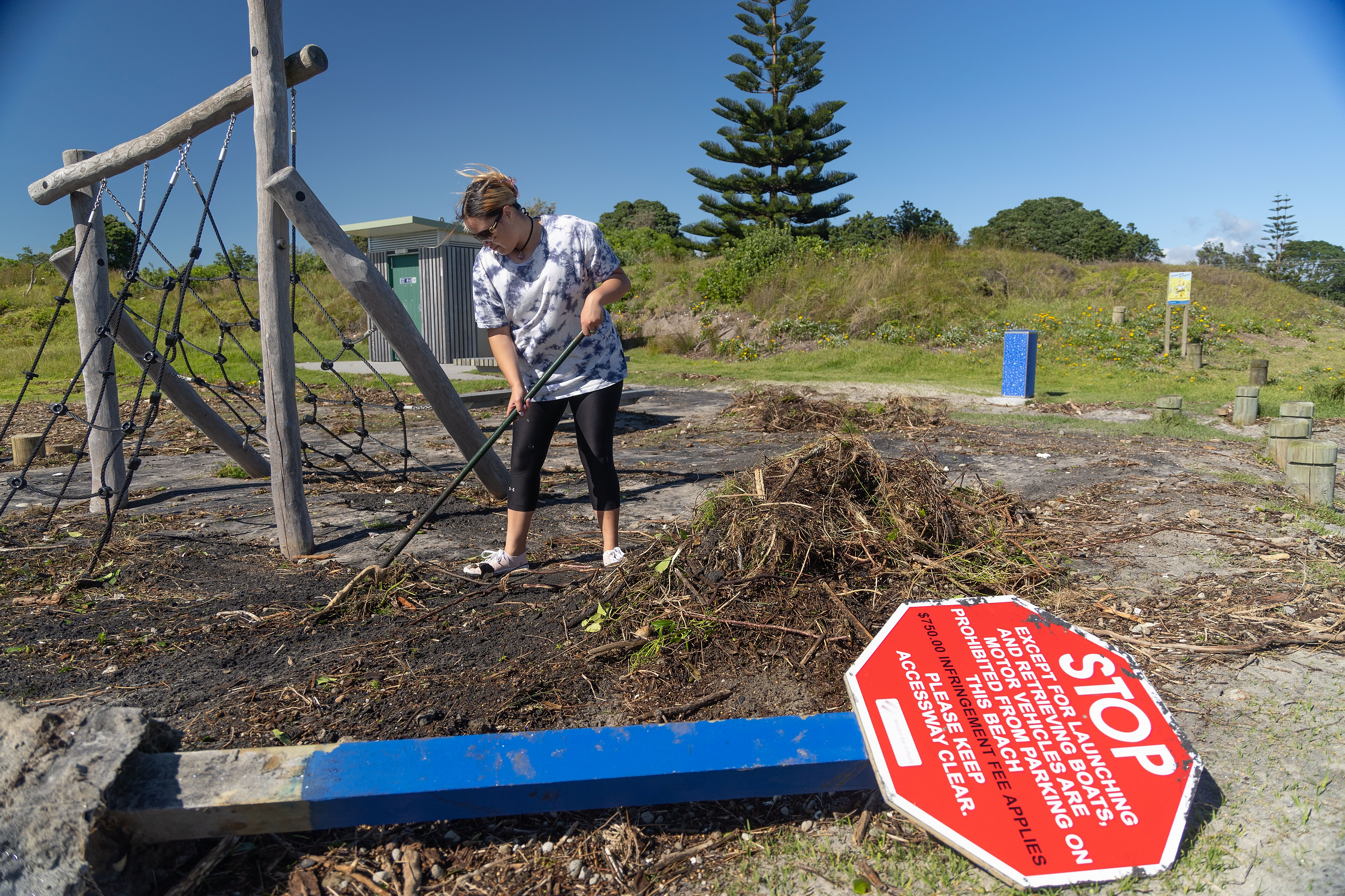 A woman in t-shirt and leggings clearing debris from a playground. There is a collapsed red sign on the ground in front and a climbing frame behind. There are piles of earth, roots and leaves.