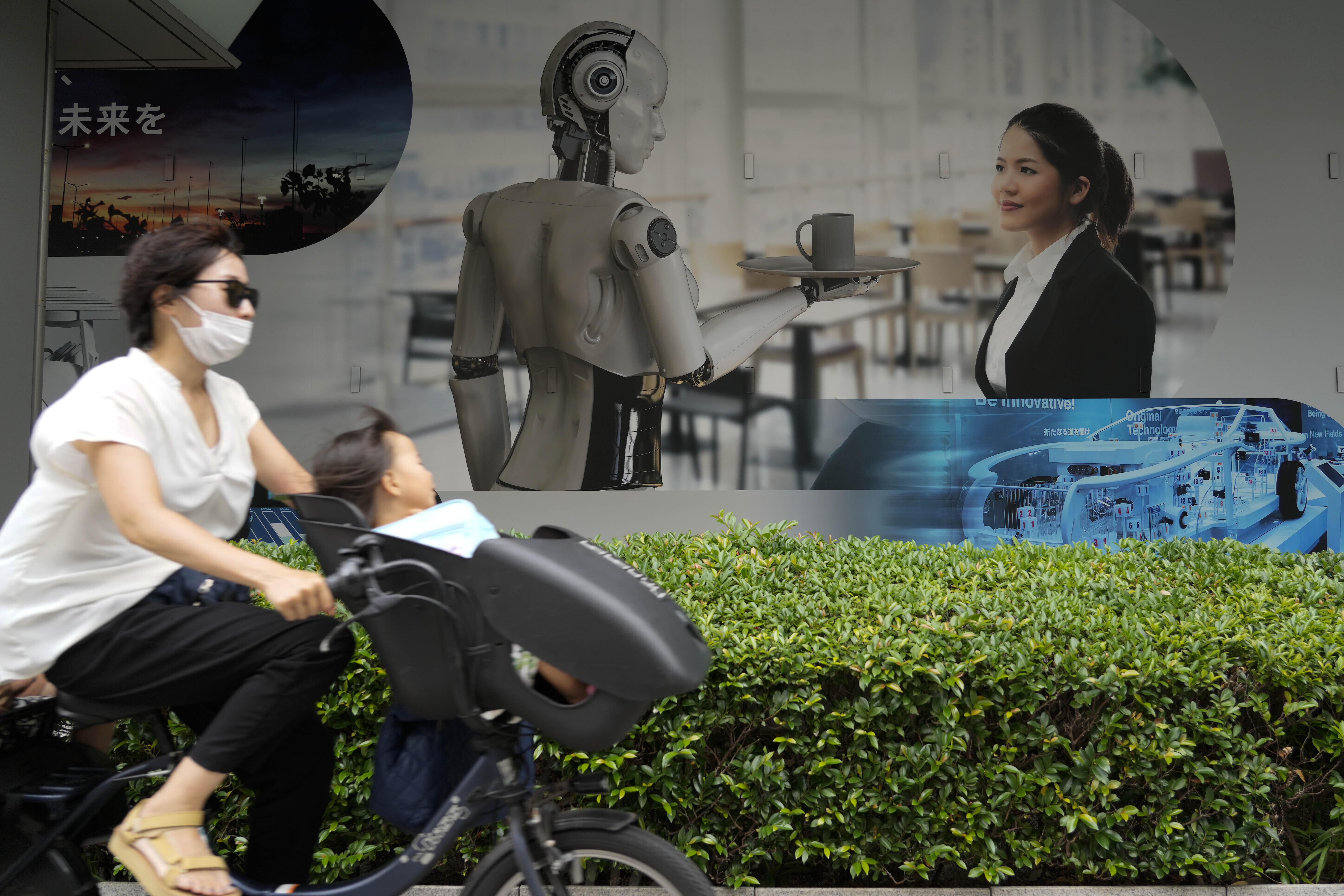A woman cycles past a billboard in Tokyo. She has a baby on the front.