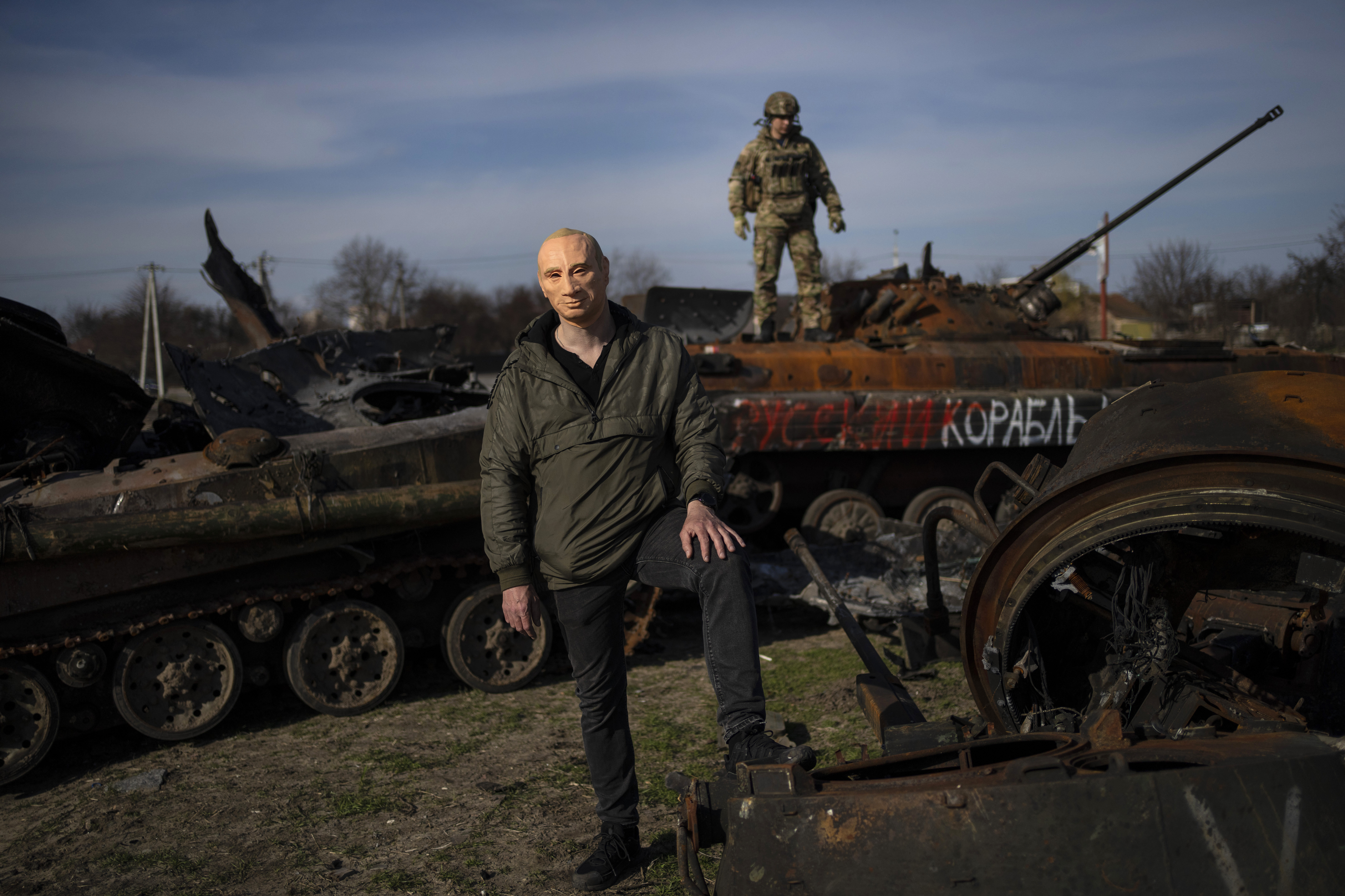 A civilian wears a Vladimir Putin mask as a spoof, while a Ukrainian soldier stands atop a destroyed Russian tank in Bucha