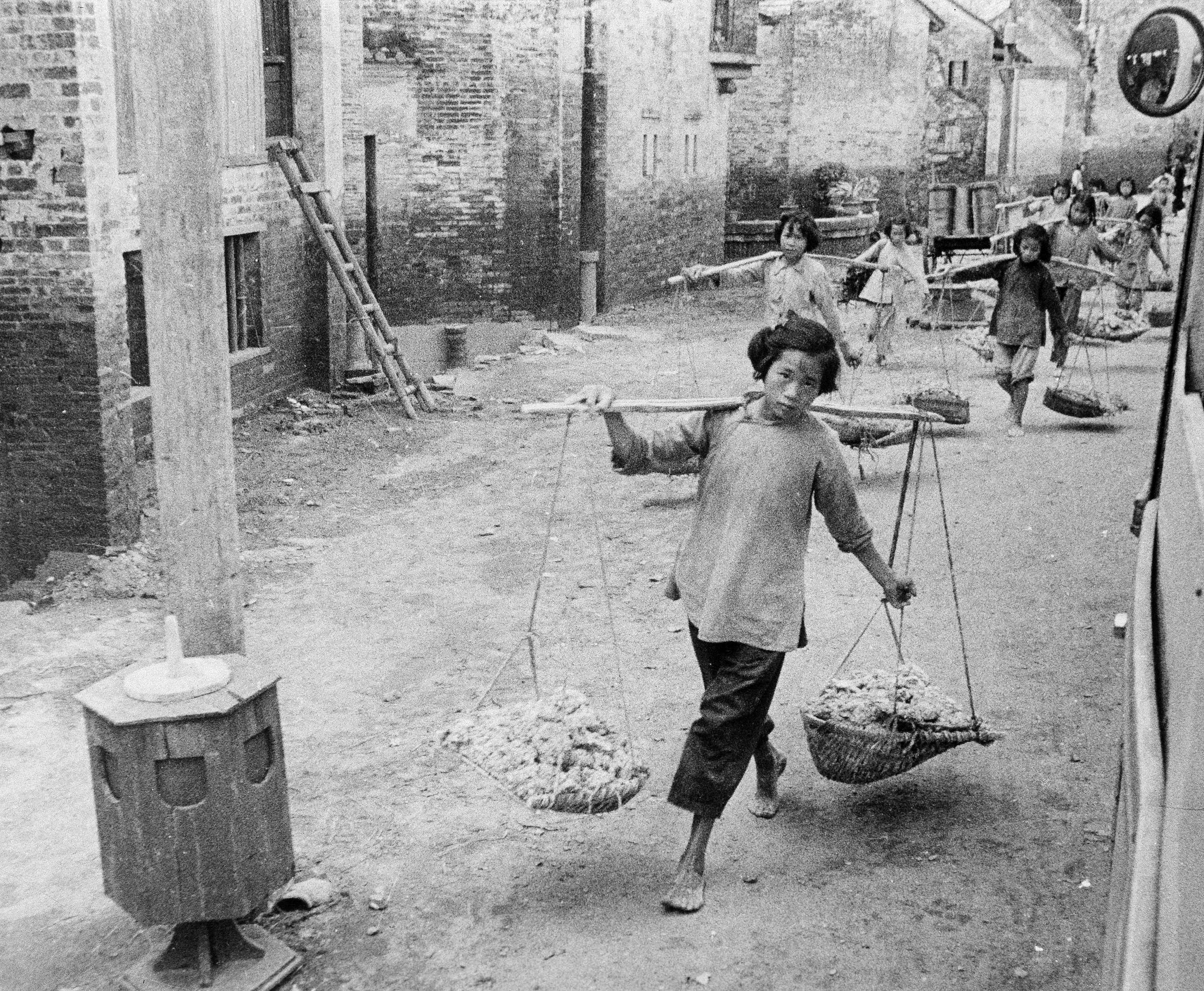 Chinese girls with yokes around their shoulders carrying what looks like bricks in the baskets hanging below. It is a black and white image from 1961.