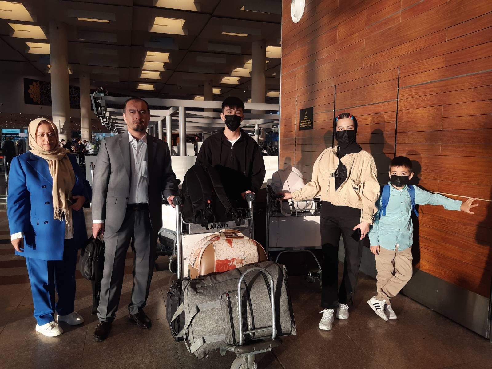 A photo of five people standing in an airport with a baggage cart with suitcases between them.