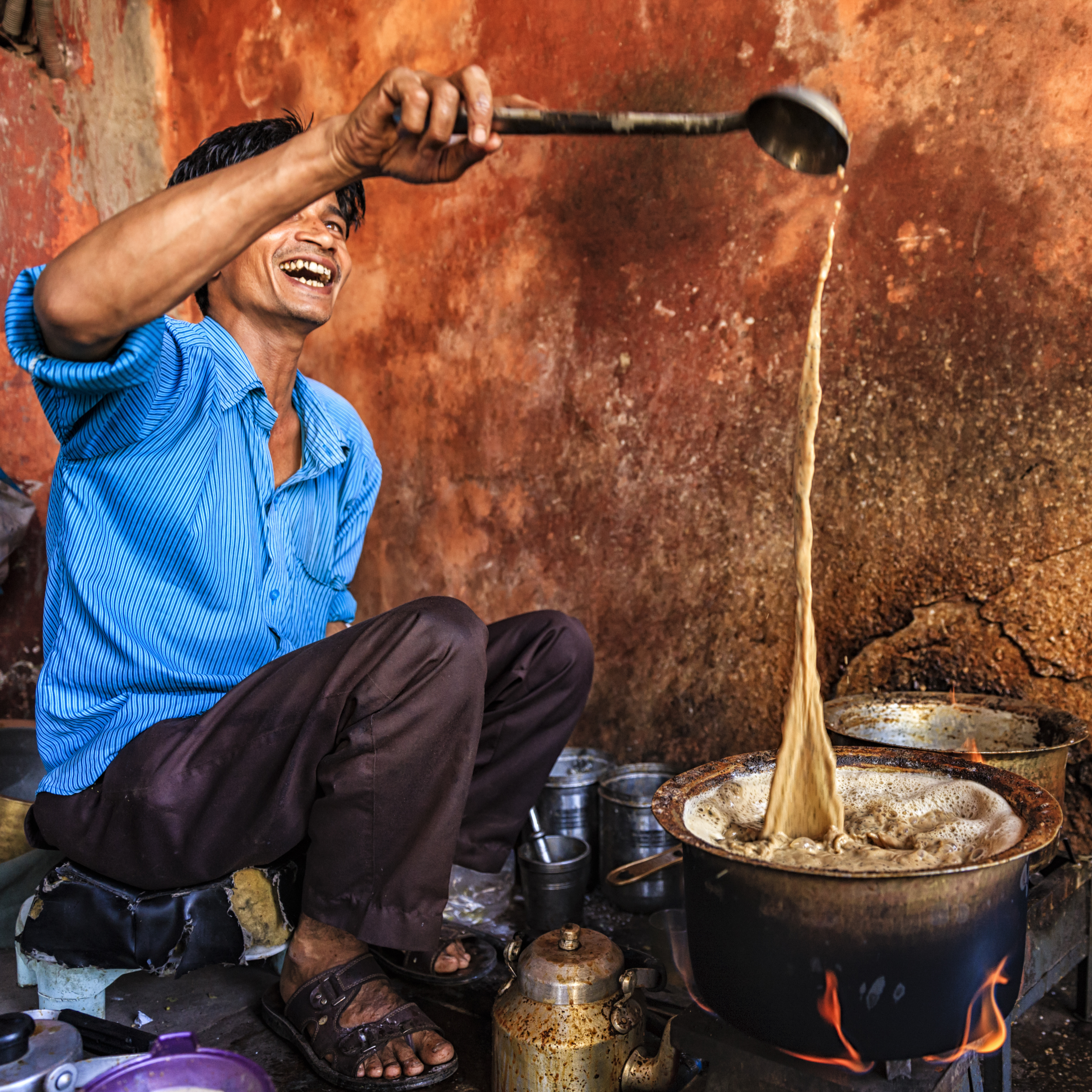 A laughing chai walla pours tea from a ladle held high back into the bubbling pot of chai