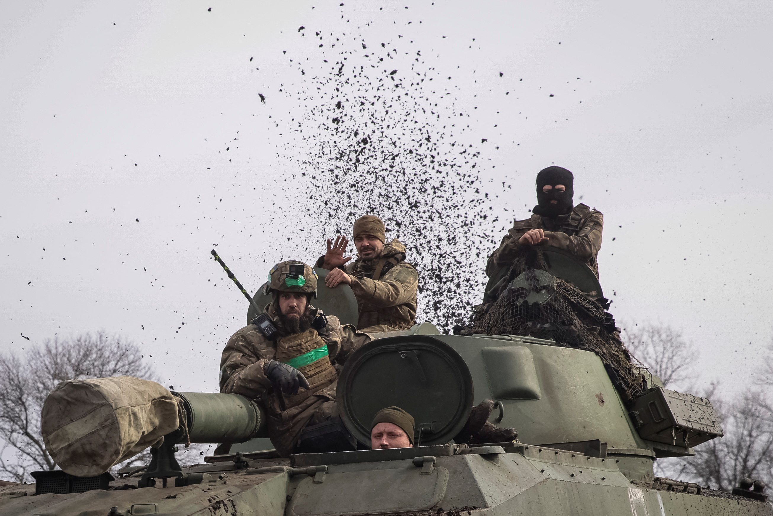 Ukrainian soldiers in a self-propelled howitzer on the frontline near Bakhmut. Three are on top of the vehicle. One is peering out of a hatch. There are clods of mud exploding into the air behind them.