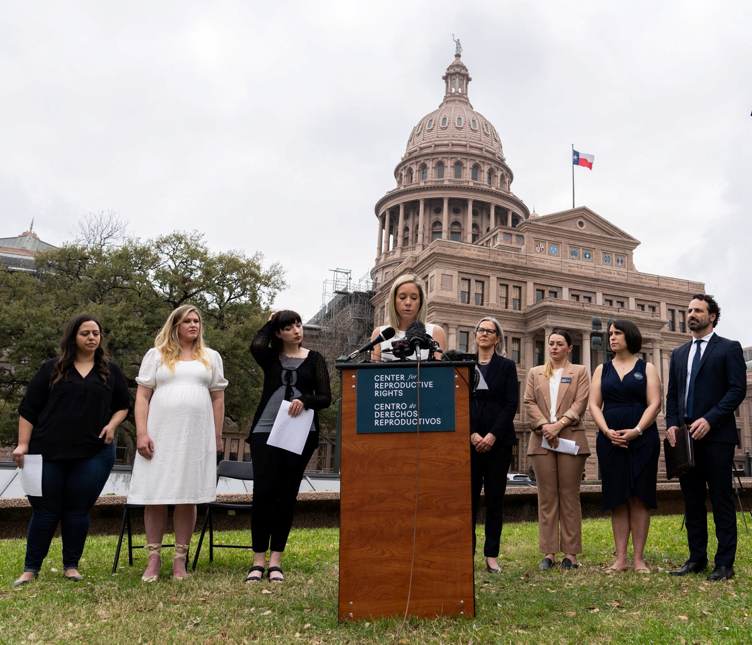 Amanda Zurawski speaks in front of the Texas State Capitol. Three women stand to her left on the grass and three to her right, with a male next to them. Amanda stands behind a pulpit that has a sign on it reading, "Center for Reproductive Rights".