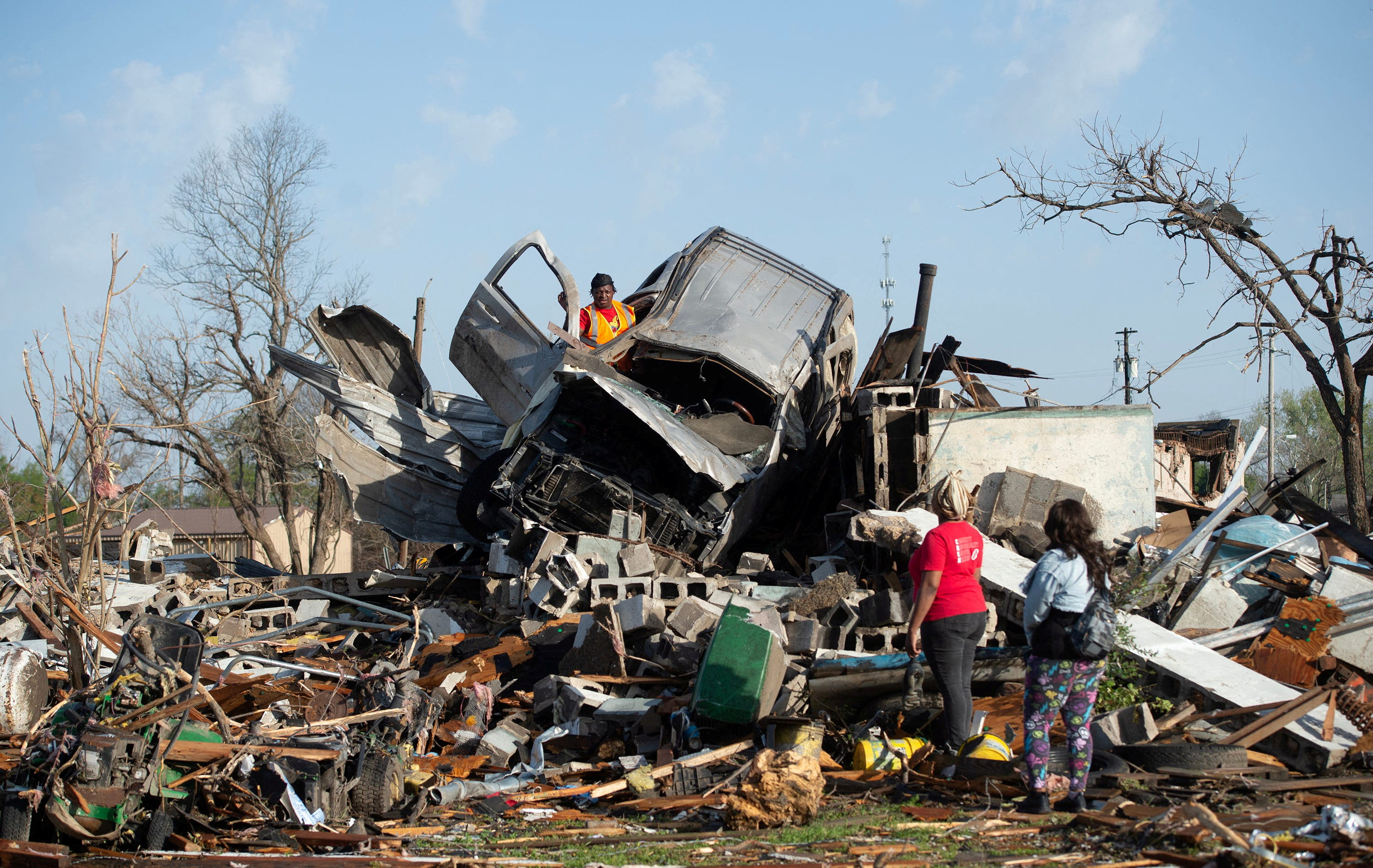 Residents of Rolling Fork, Mississippi, survey damage from a tornado. A visibly-damaged car, possibly a SUV, is lying on top of a mound of debris.