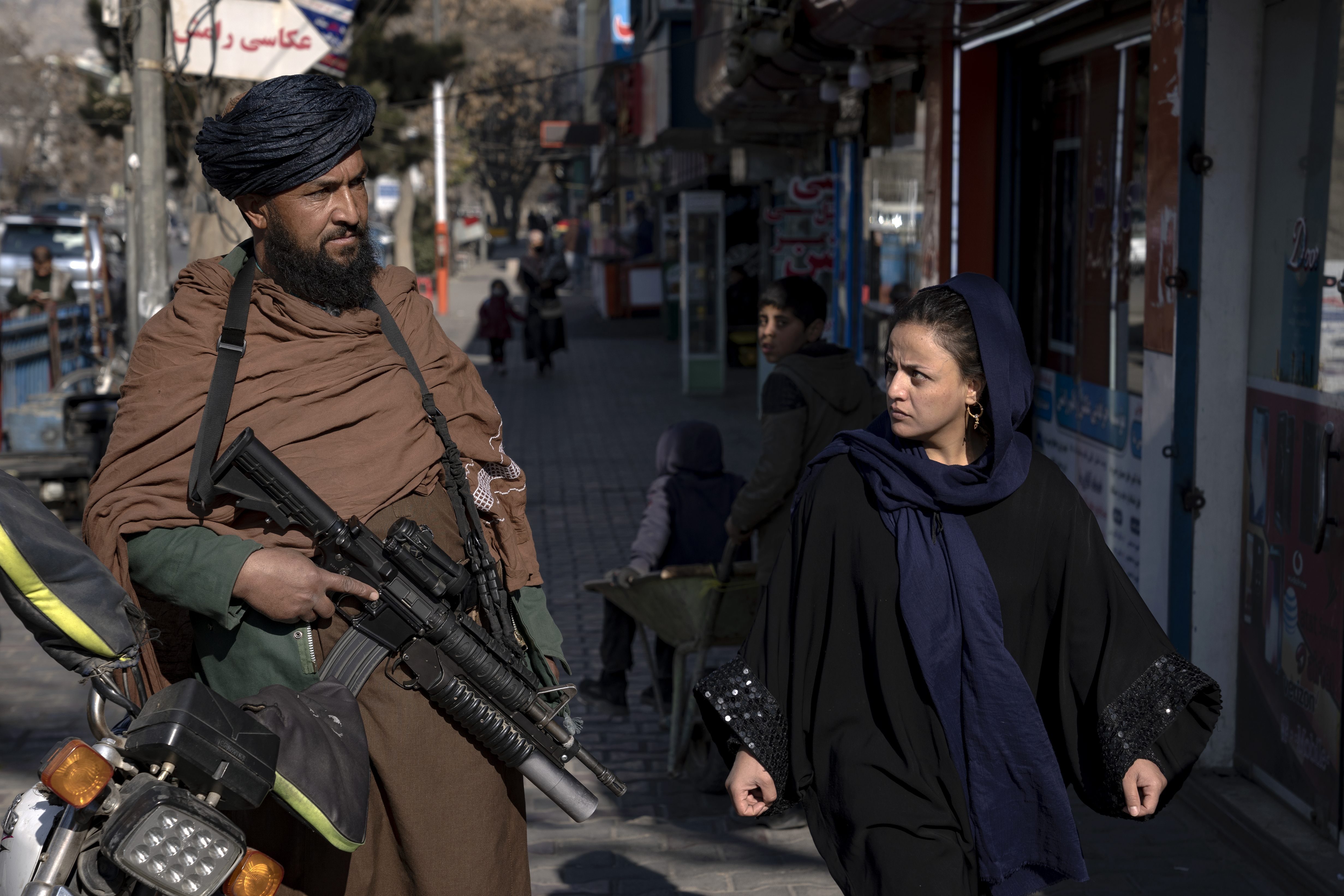 A Taliban guard stands guard as a woman walks past in Kabul, Afghanistan, on December 26, 2022 [Ebrahim Noroozi / AP Photo]