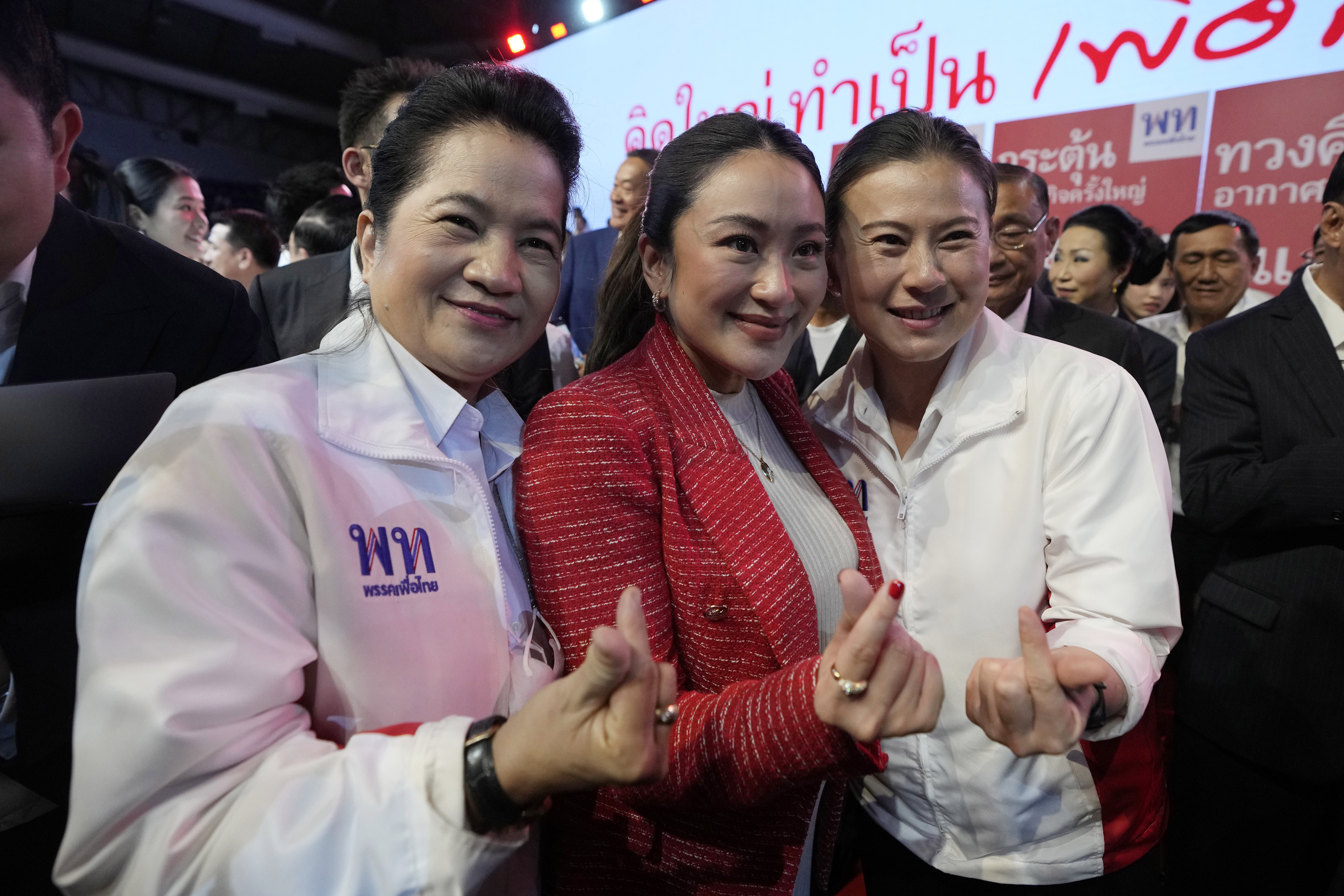 Paetongtarn Shinawatra with two supporters at a Pheu Thai rally. She's wearing a red jacket and is smiling. They are making the mini heart gesture with their fingers.