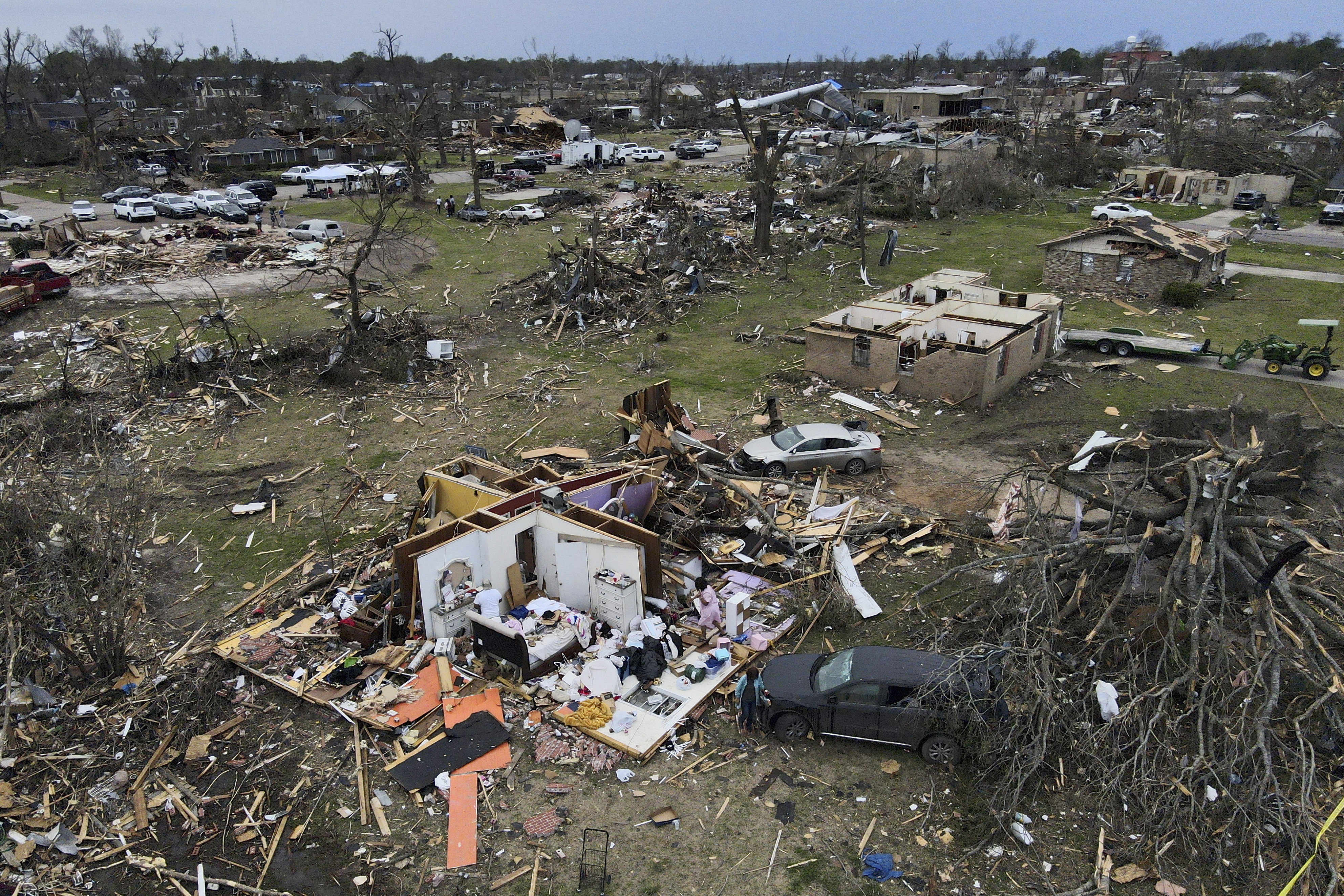 An aerial view of a neighbourhood destroyed shows a house where the roof and outer walls are missing. There is debris everywhere as well as trees stripped of leaves lying on the ground. 