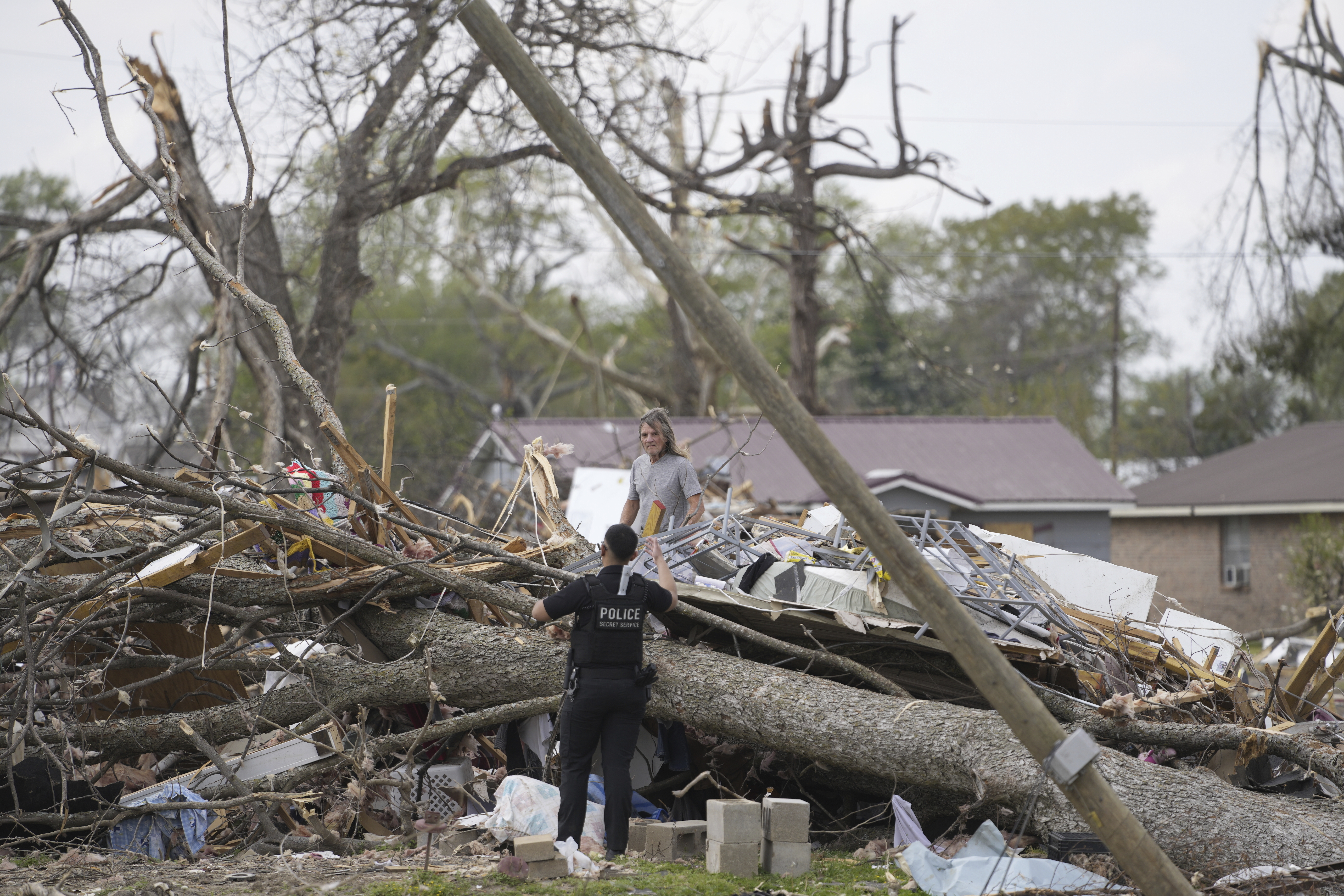 The police officer is talking to a person on the other side of a large fallen tree. There looks to be a telegraph pole leaning to the side.