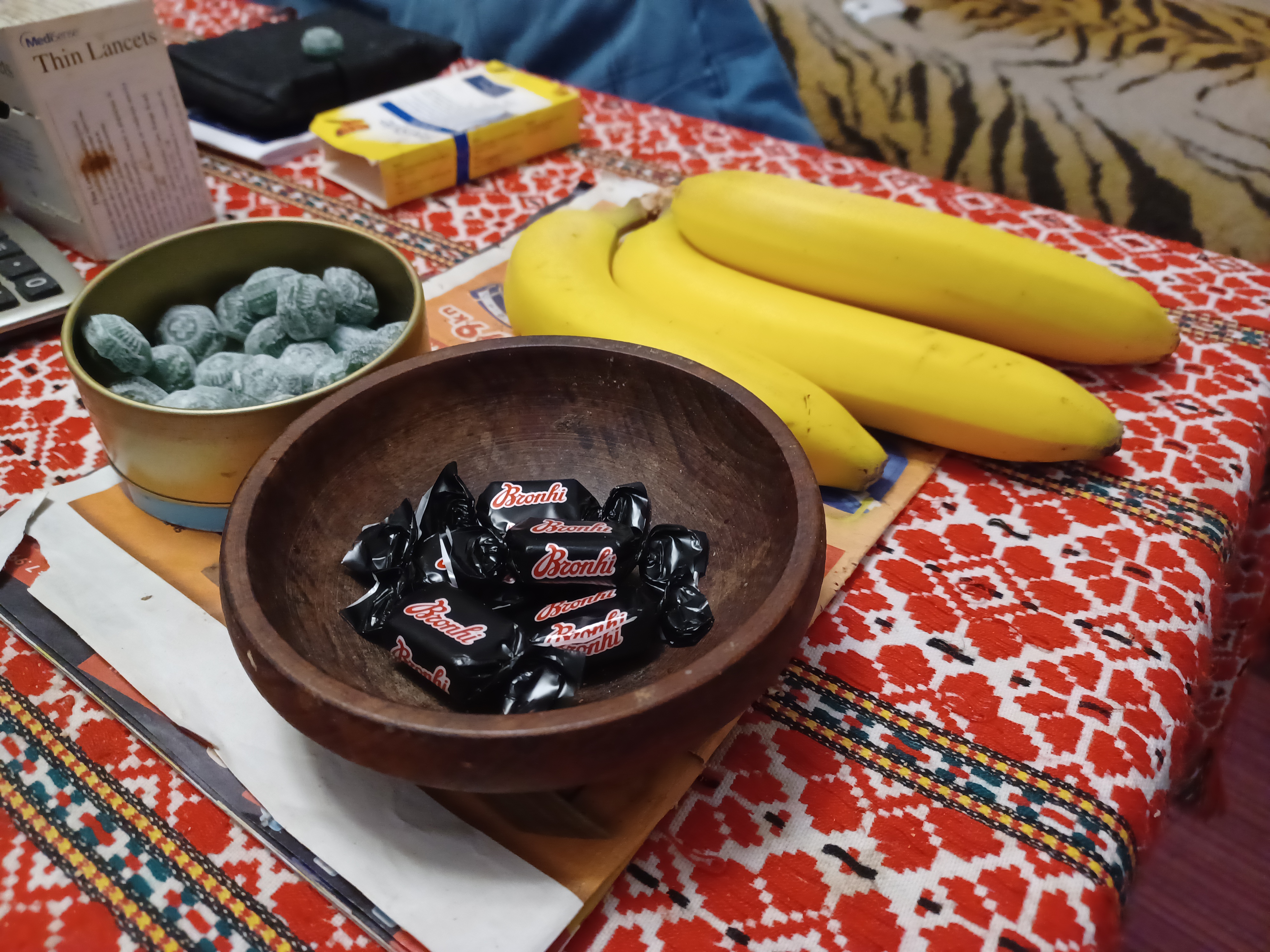 A photo of individually wrapped sweets in a bowl with a bunch of bananas on one of the sides and a bowl of exposed sweets on one of the the other sides..