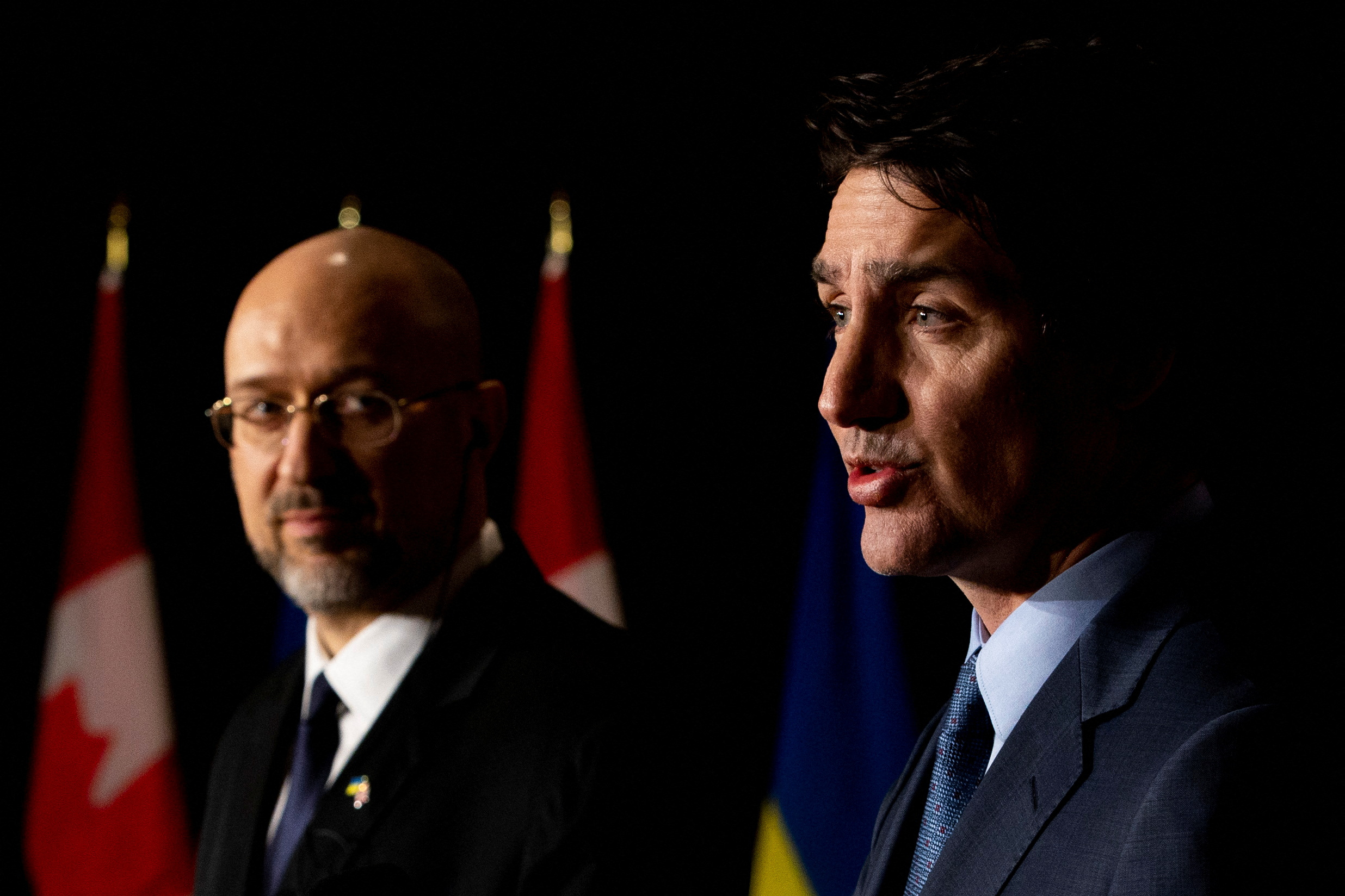 In a darkened room with flags, a man in glasses looks at Justin Trudeau as he speaks