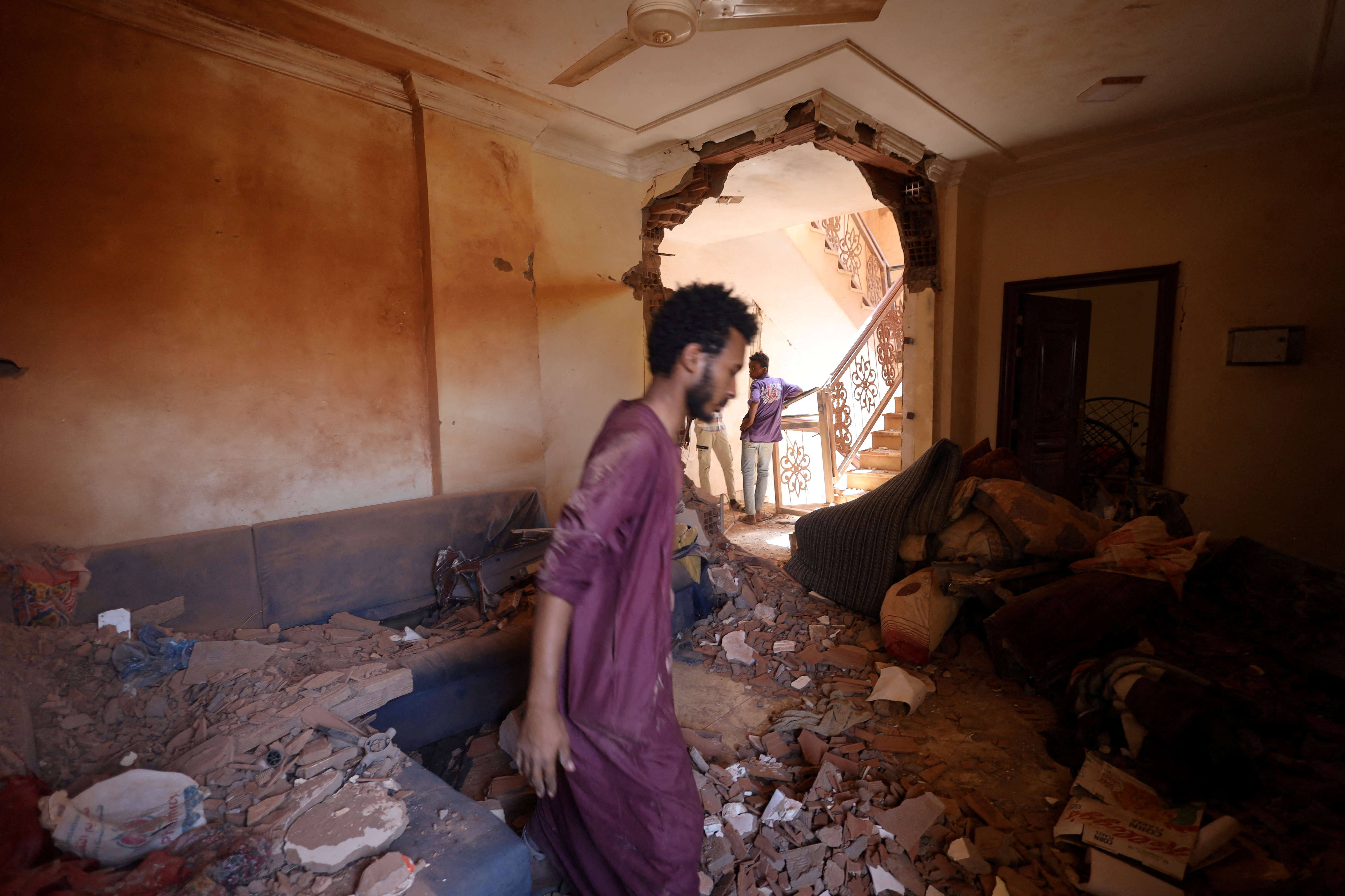 A man stands amid rubble in a Khartoum house hit during fighting between the army and RSF, The doorway behind hin is damaged, There is rubble all over the floor and one the large sofas against two walls