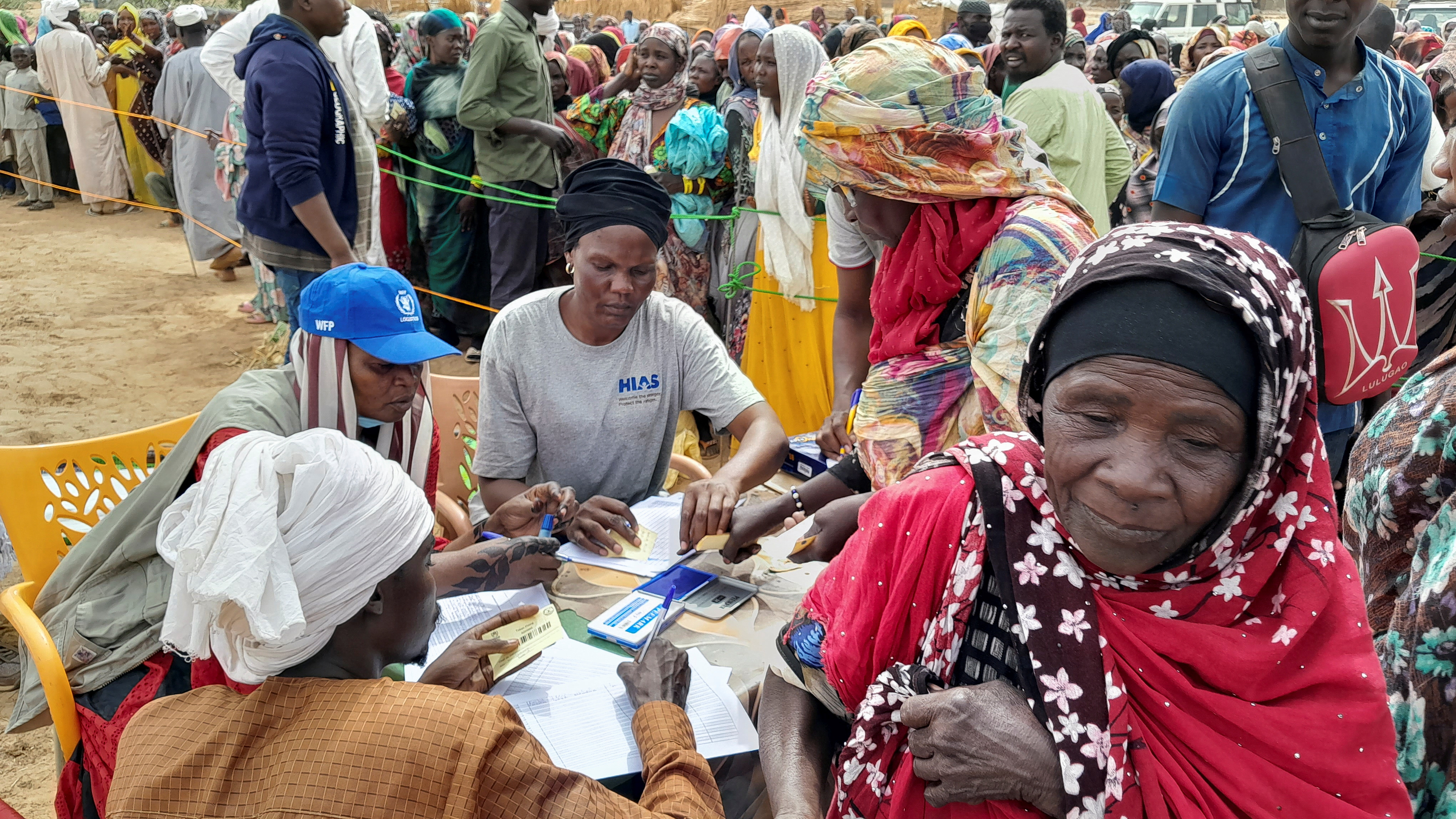 Women getting supplements at a WFP registration desk after fleeing Sudan. They are outside. The woman at the desk has lots of pieces of paper in front of her.