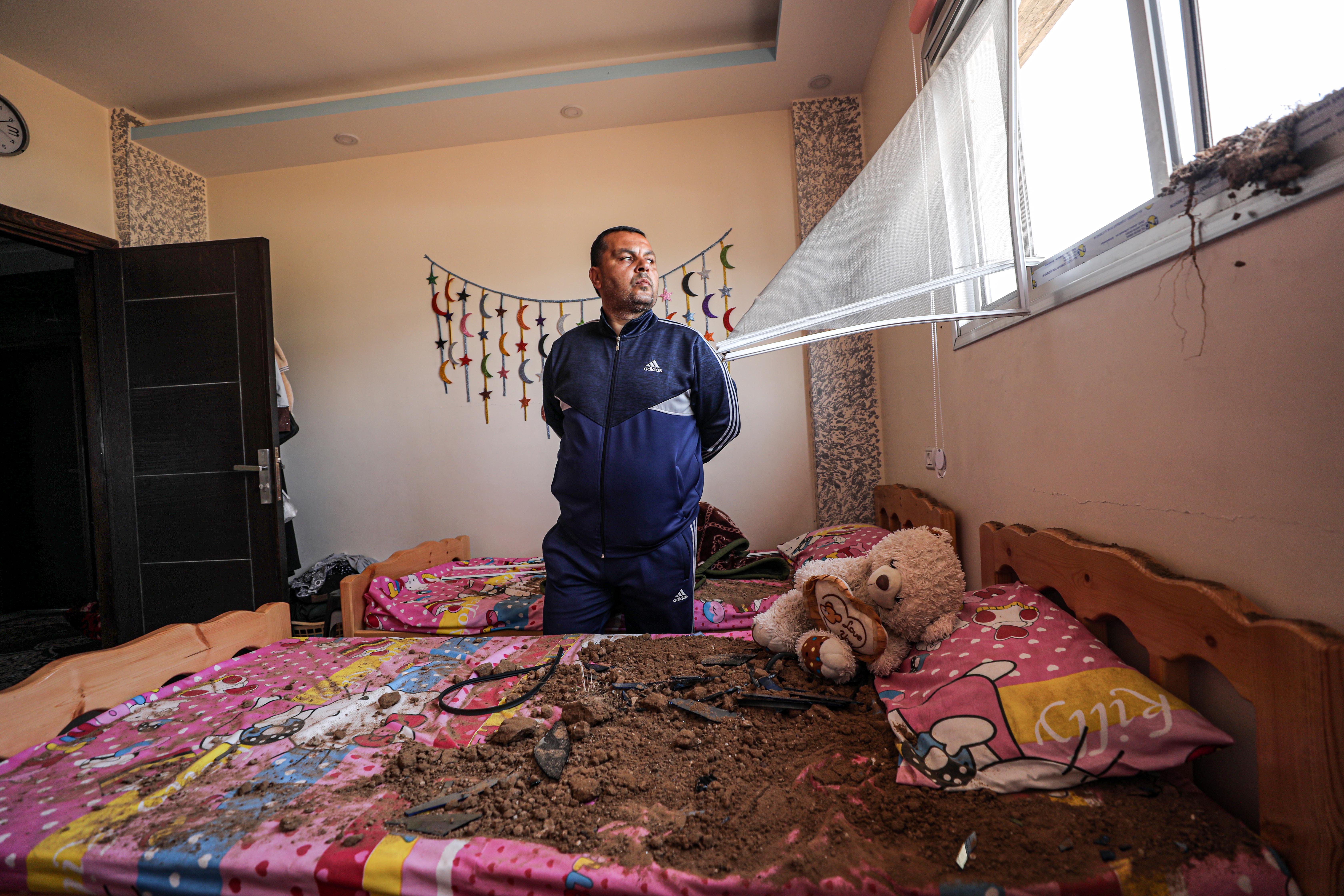 The father stands in between two beds. Both bends have pink and yellow bedcovers. The bed in front of him is covered with dirt on which lies a pink pillow and teddy bear. He is looking out the window, which has been damaged. The fly screen is bent and protrudes towards him.