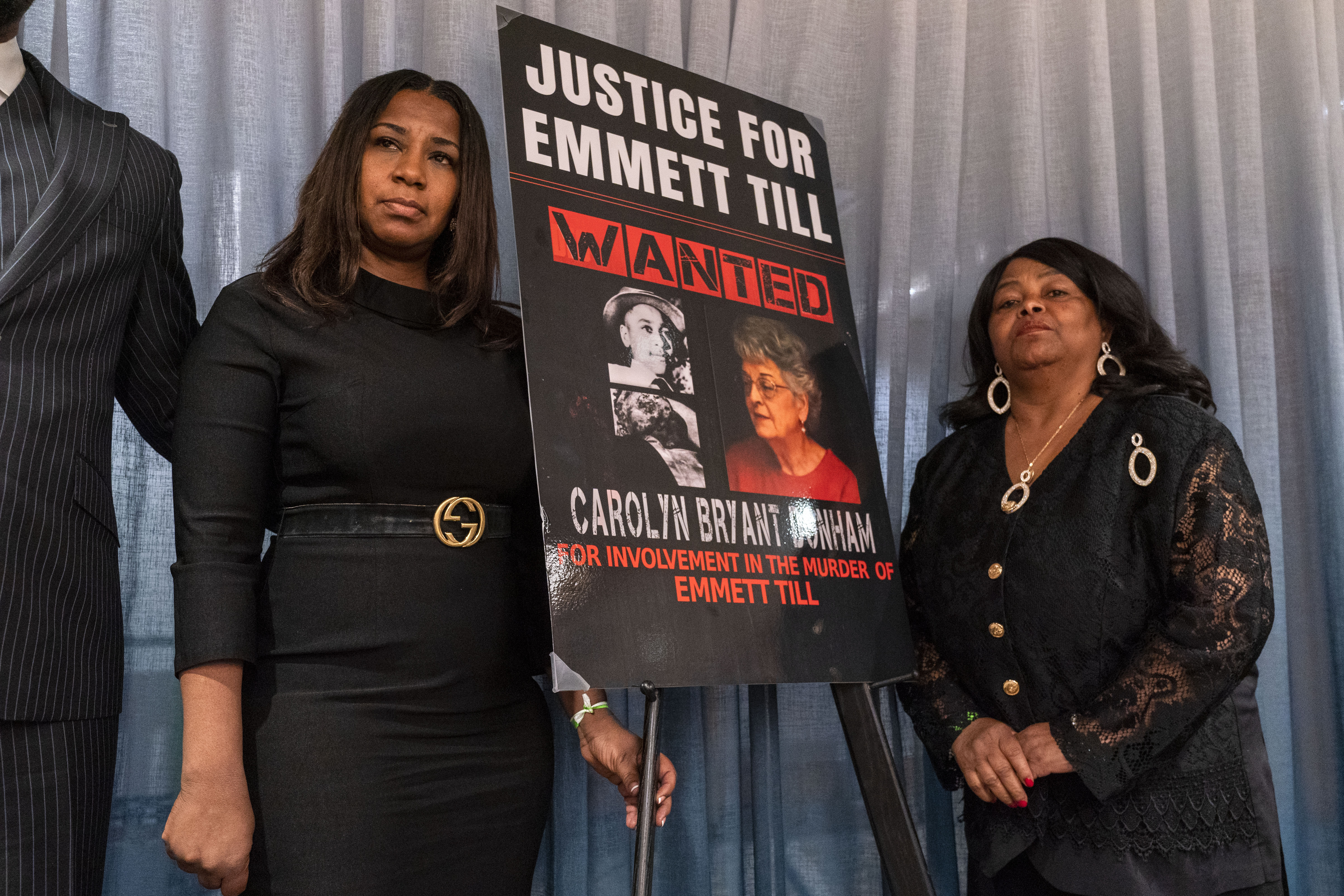 Two women stand on either side of a sign that reads: "Justice for Emmett Till. Wanted: Carolyn Bryant Donham"