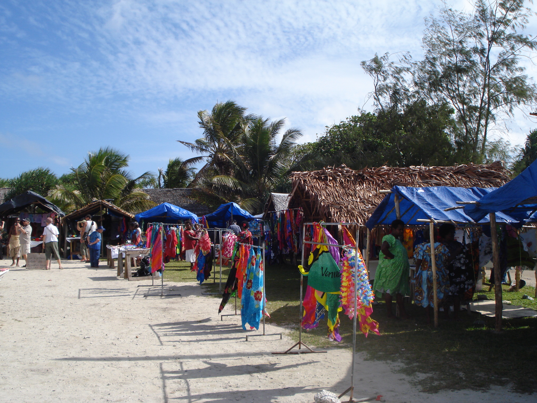 Stalls along a beach in Vanuatu. The stalls are mostly palm thatch and are selling colourful clothing. There are palm trees behind