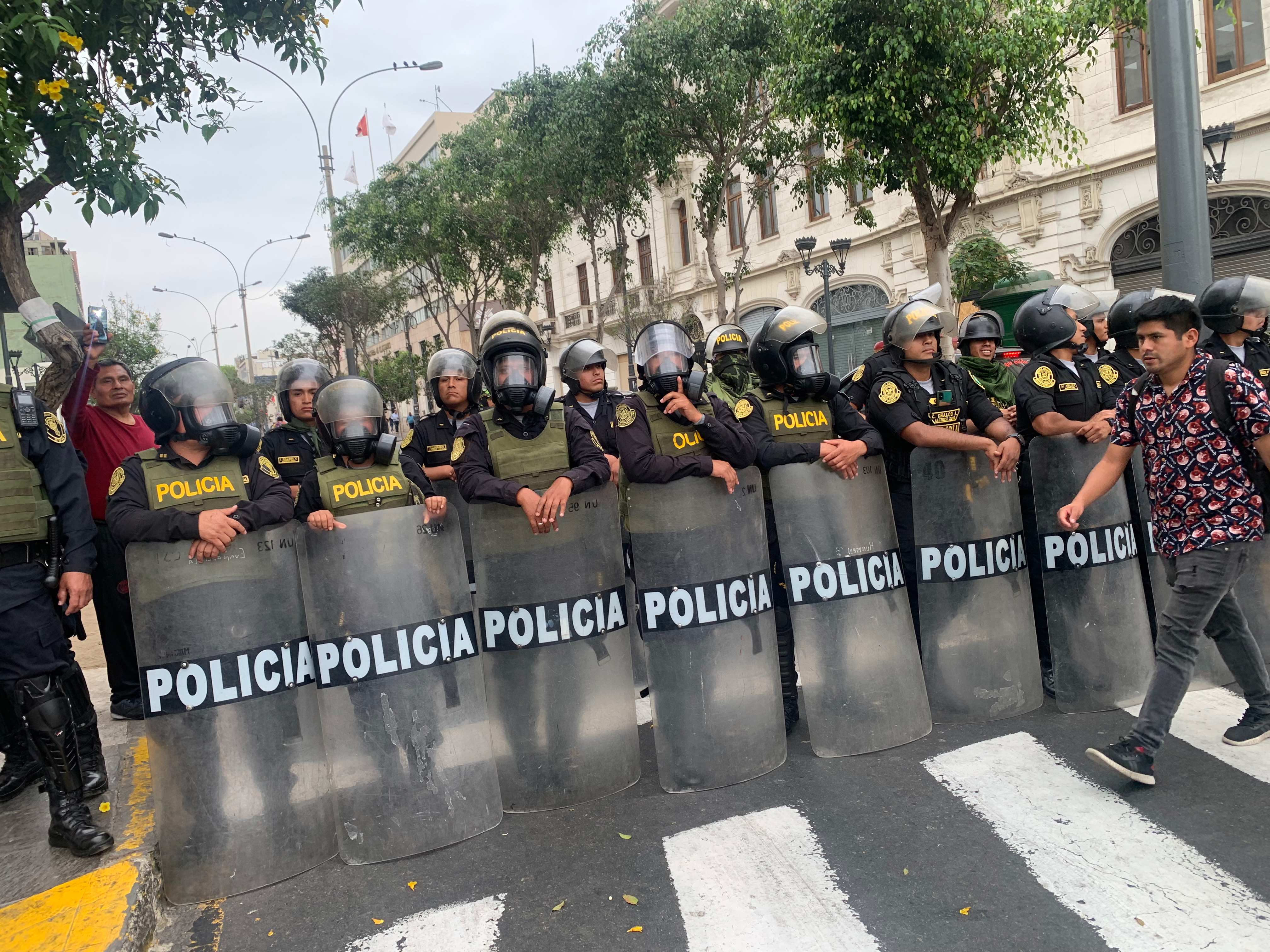 A man crosses a street blocked by a line of police officers in riot gear carrying plastic shields labeled “policia”