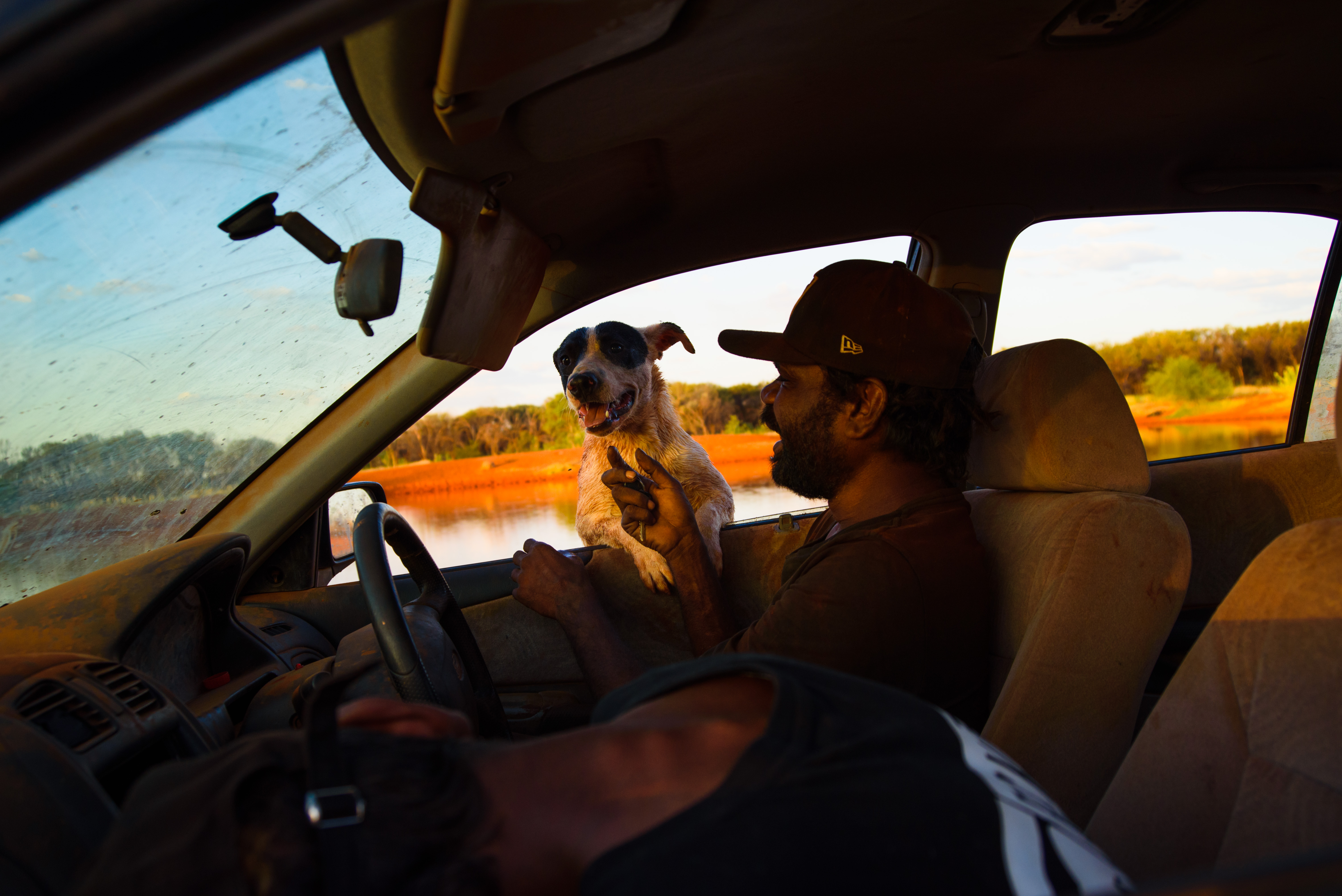Lloyd Jampijinpa Brown sitting on the driver's side of a car. He is petting his dog Blackeye which is leaning in to the car from outside, its paws resting on the windowsill.