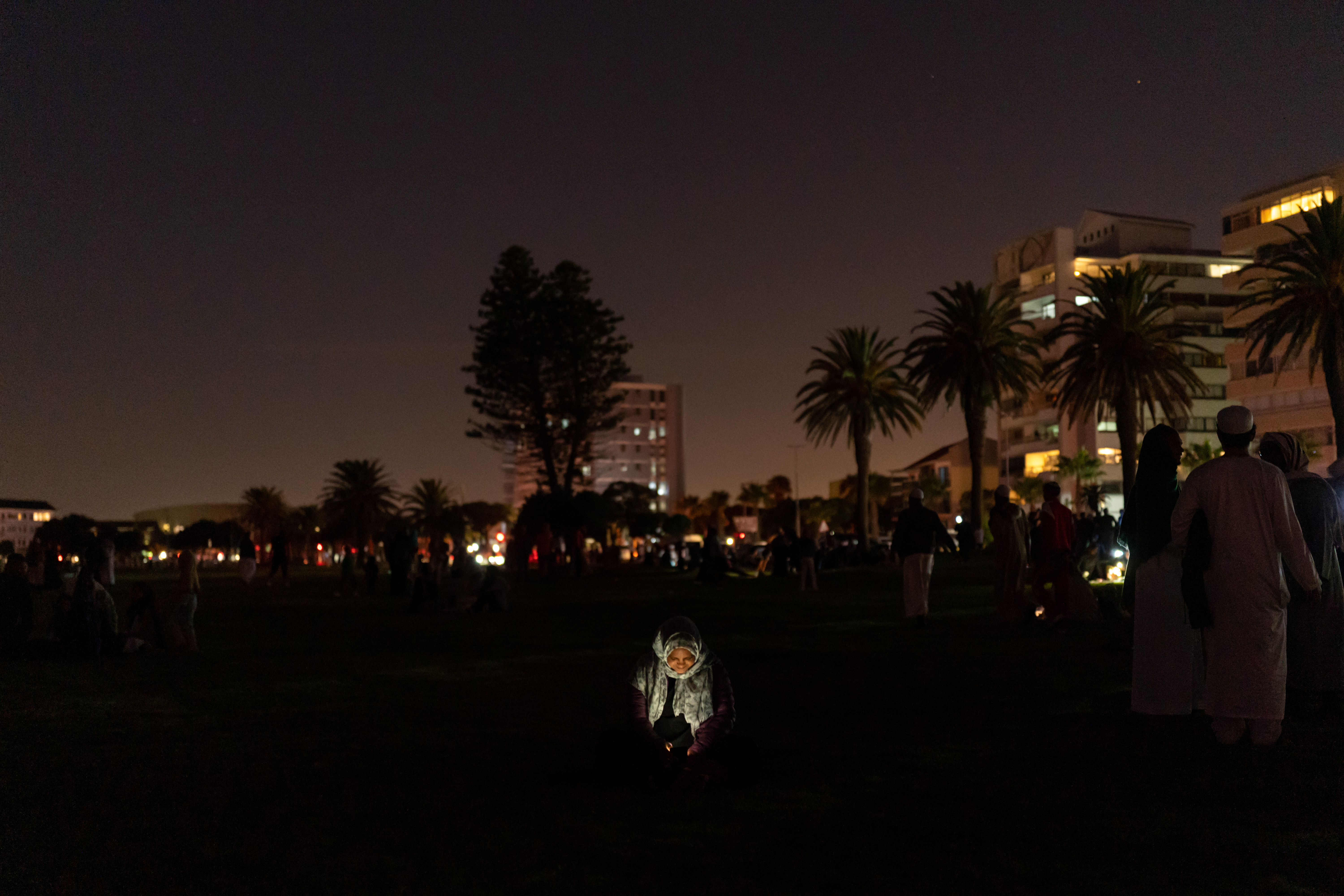A photo of a person in the dark with people, trees and buildings in the background.