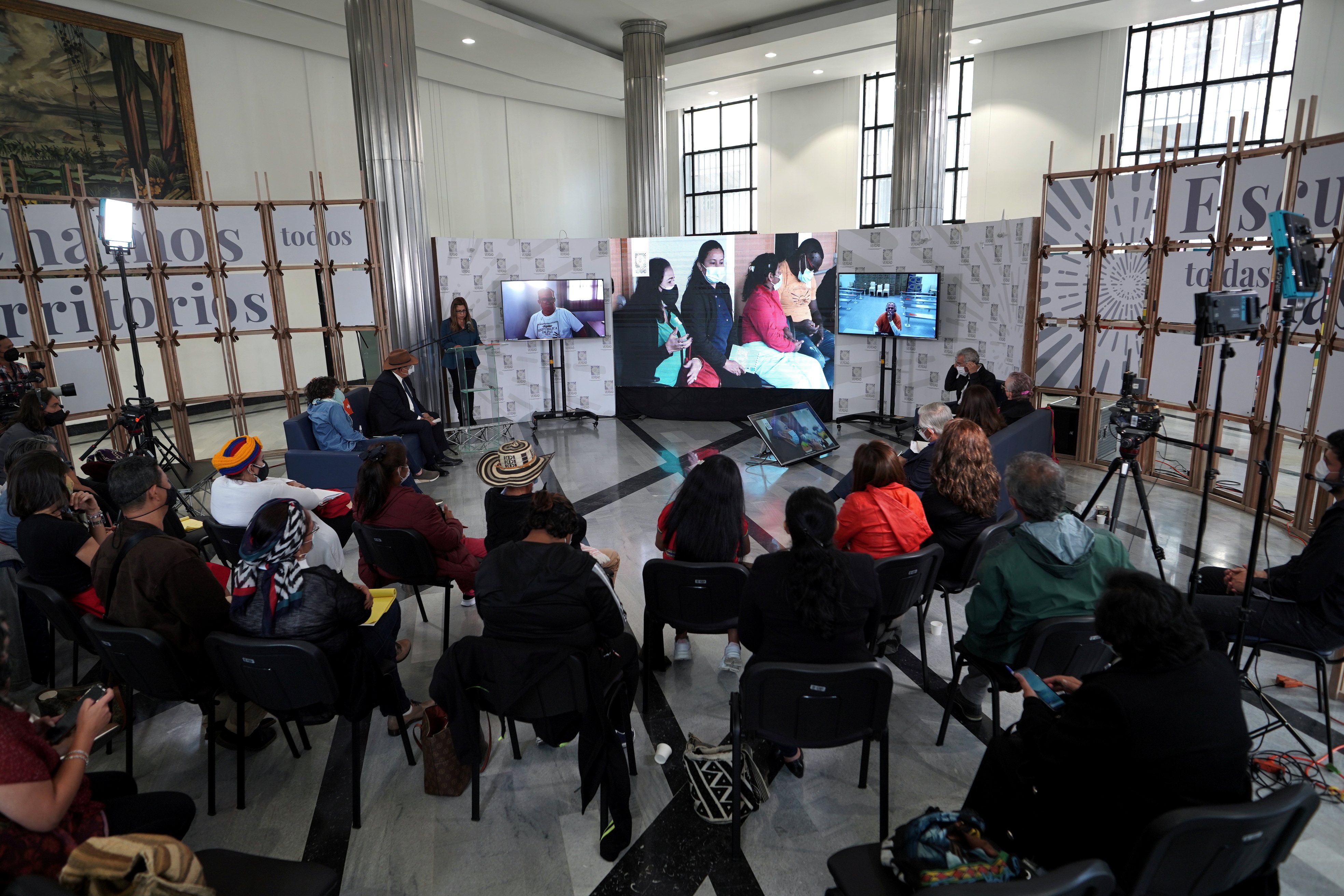 People gather around a set of TV screens in an indoor setting, where speakers can be seen.