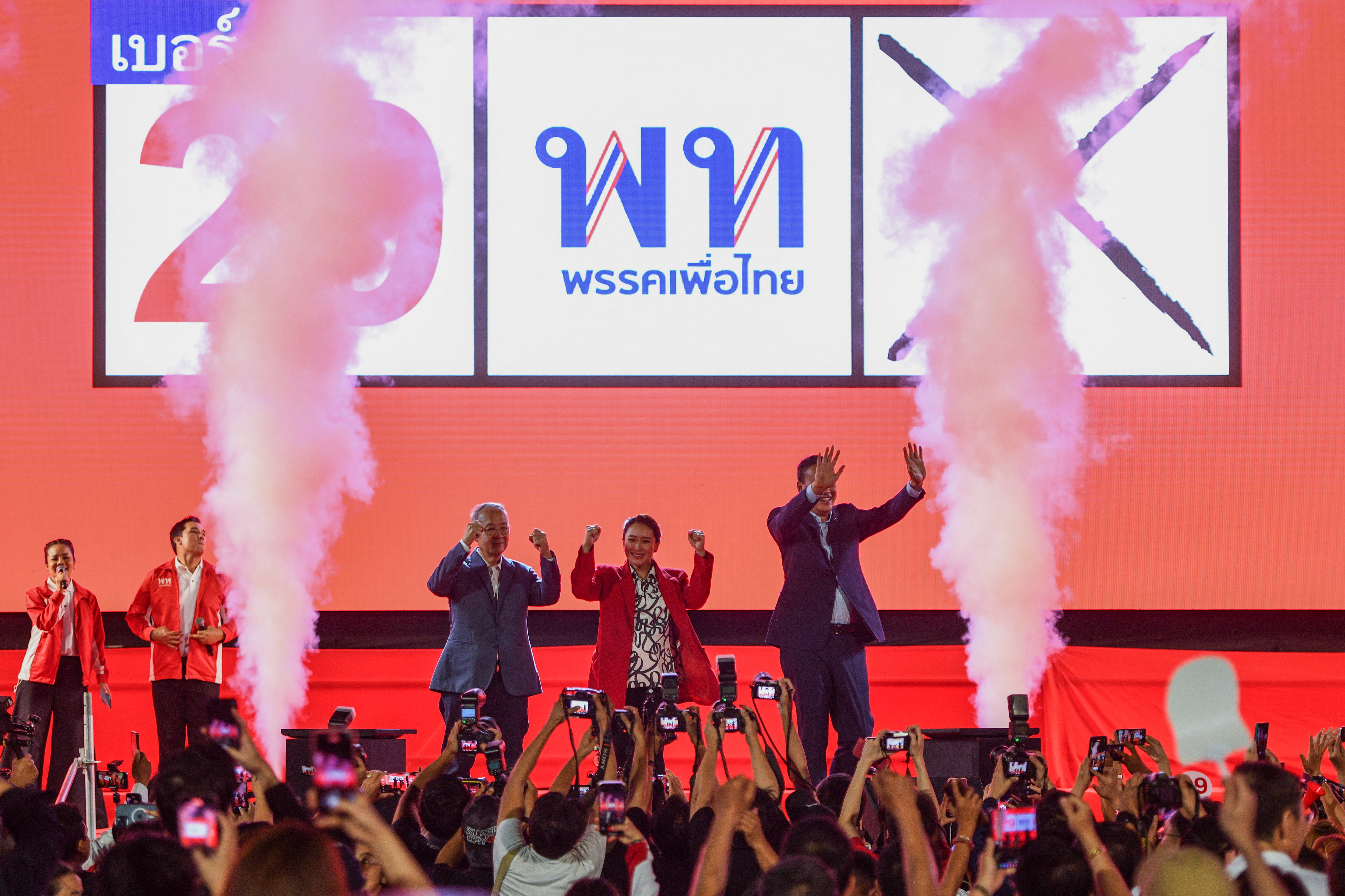 Paetongtarn Shinawatra as she was announced Pheu Thai's prime minister candidate. She is on stage, against a red backdrop and party logo. There are pink flares on either side of her. The crowd are clapping.