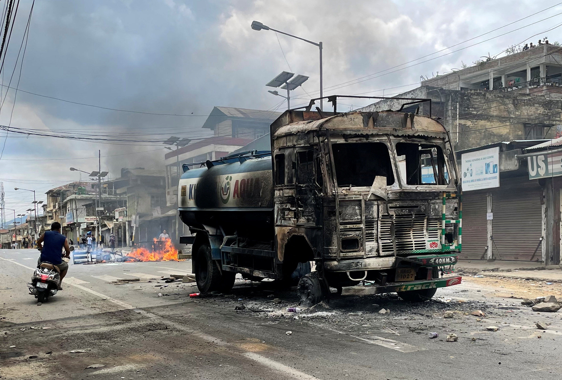 A scooterist rides past a damaged water tanker that was set afire during a protest by tribal groups in Churachandpur district.