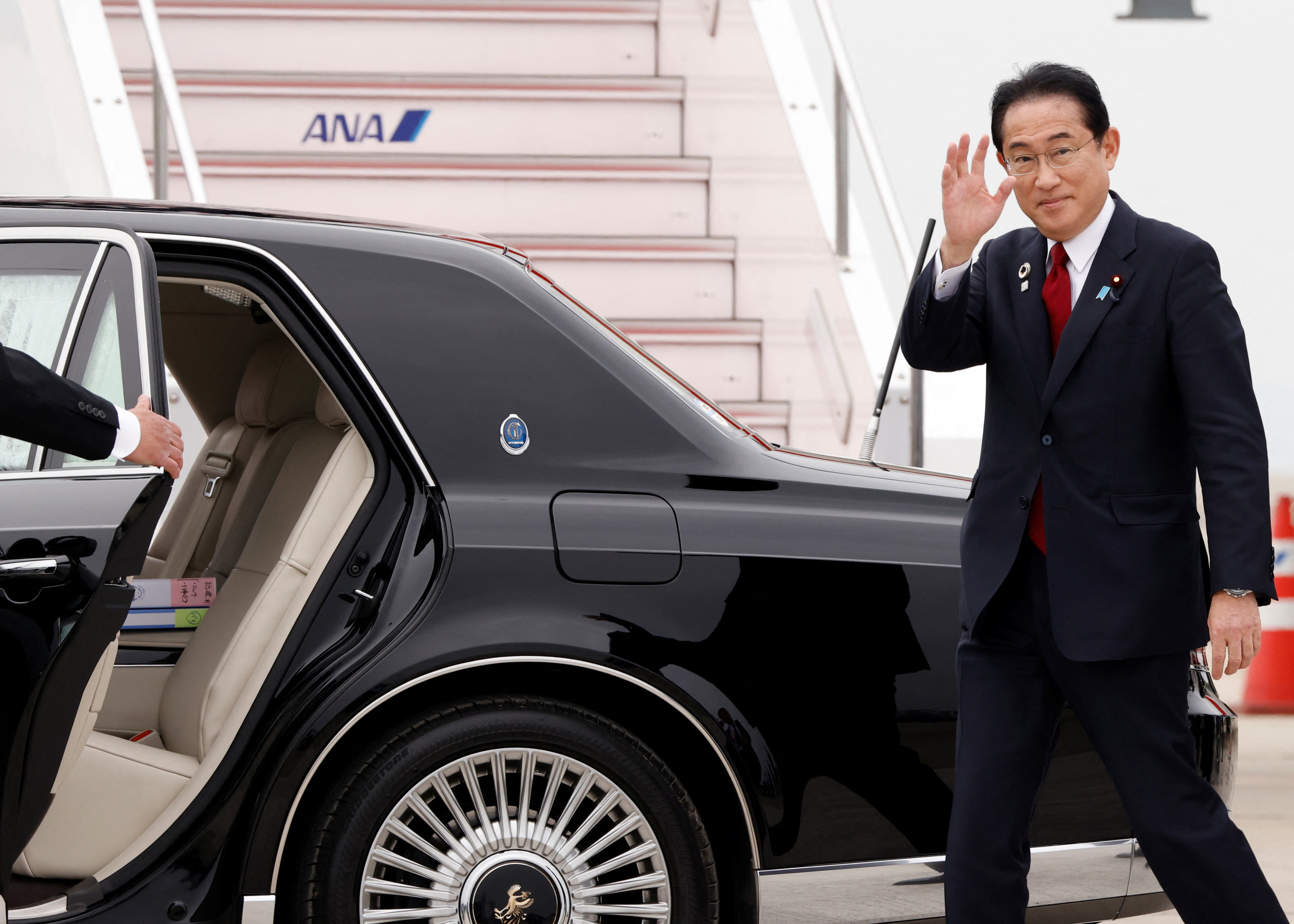 Japanese PM Fumio Kishida waves as he walks to a waiting black limousine after getting off a plane at Hiroshima airport, He is smiling.