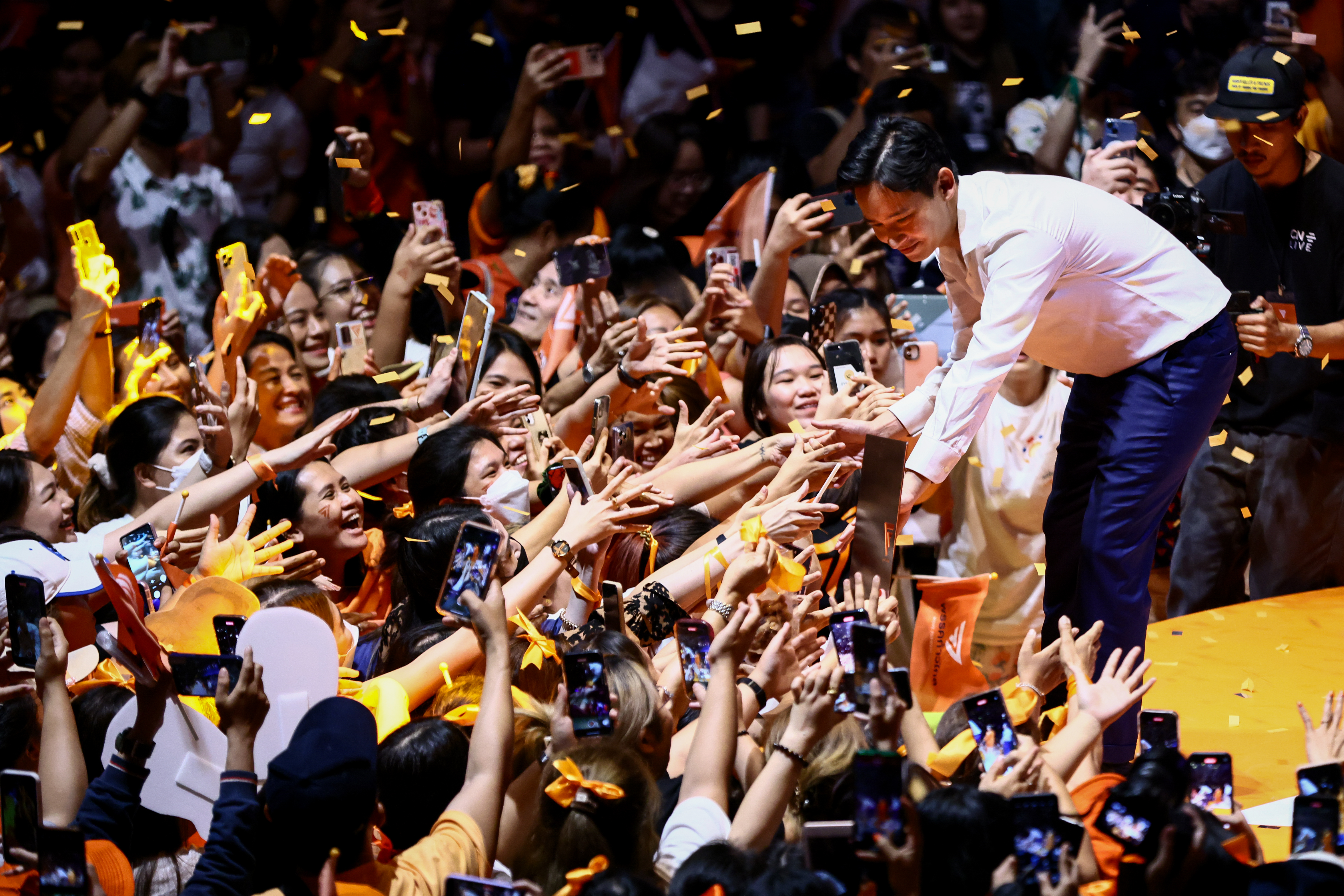 Supporters reaching out to Move Forward party leader Pita Limjaroenrat. He is standing on the stage bending down and shaking their hands.