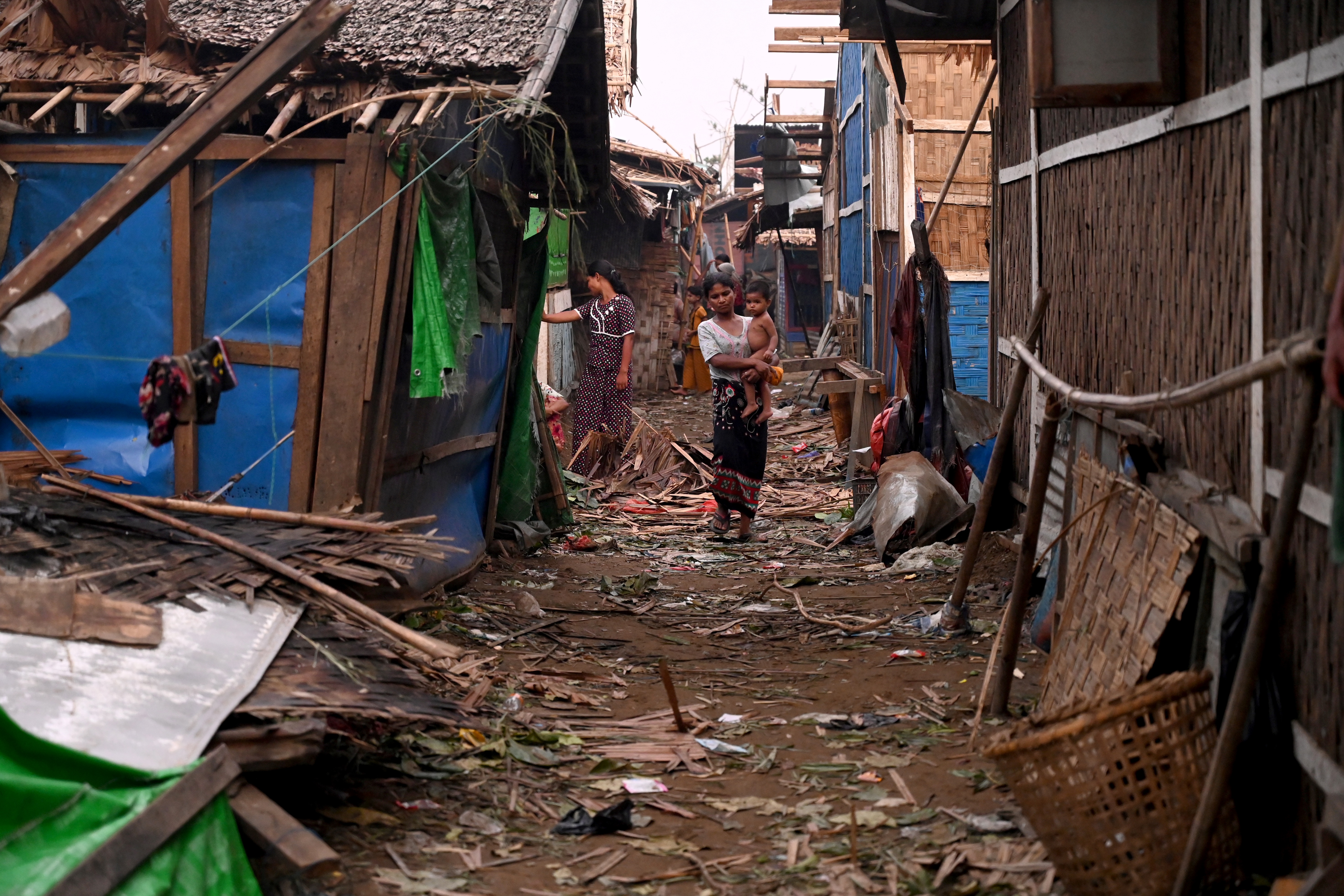 A woman walking through a debris strewn alleyway between bamboo and tarpaulin houses in a Rohingya camp in Sittwe. She is holding a child. There is another woman behind her looking inside a building