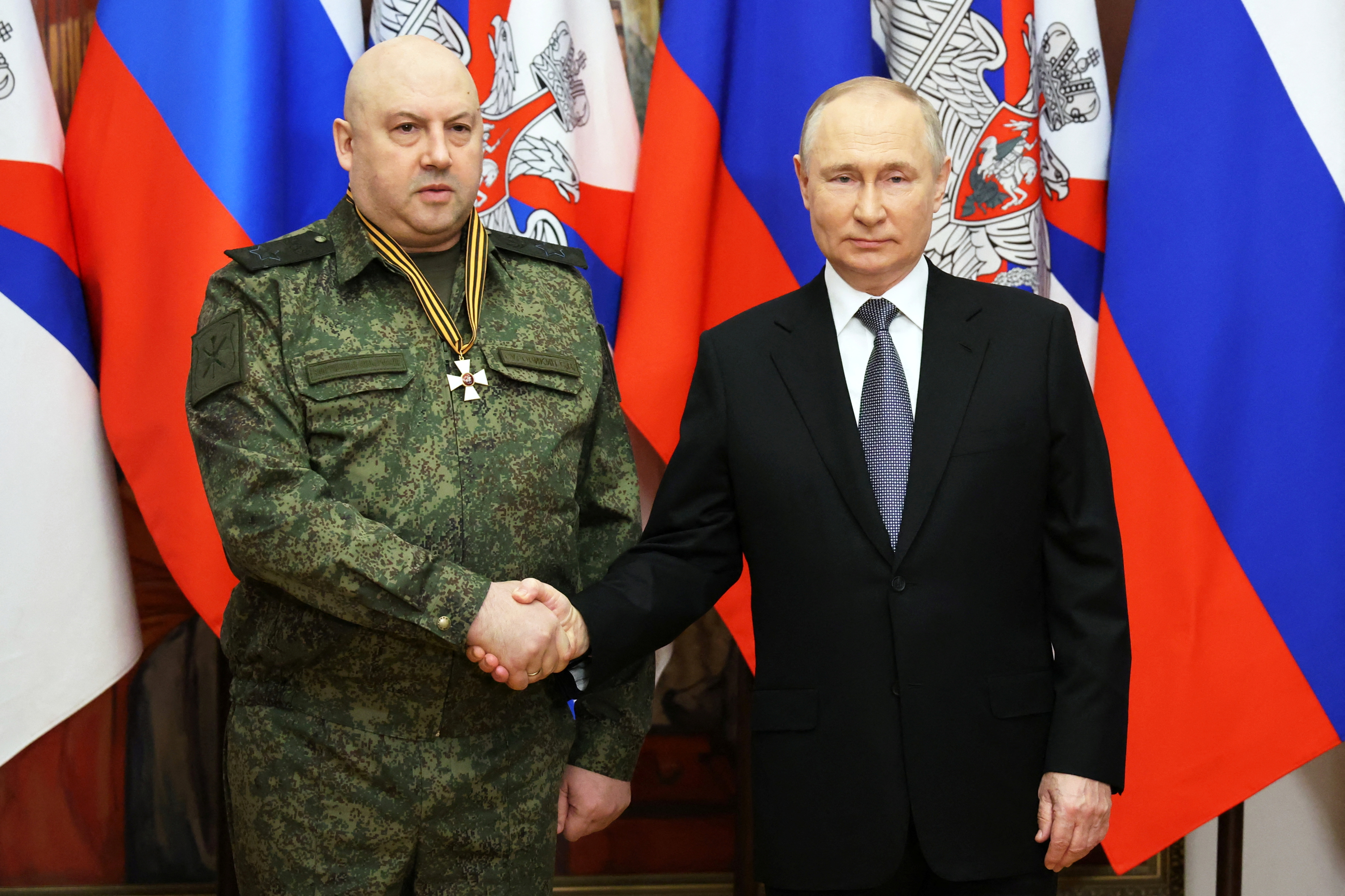 A man in military fatigues with a medal around his neck shakes hands with Vladimir Putin, who wears a suit and tie