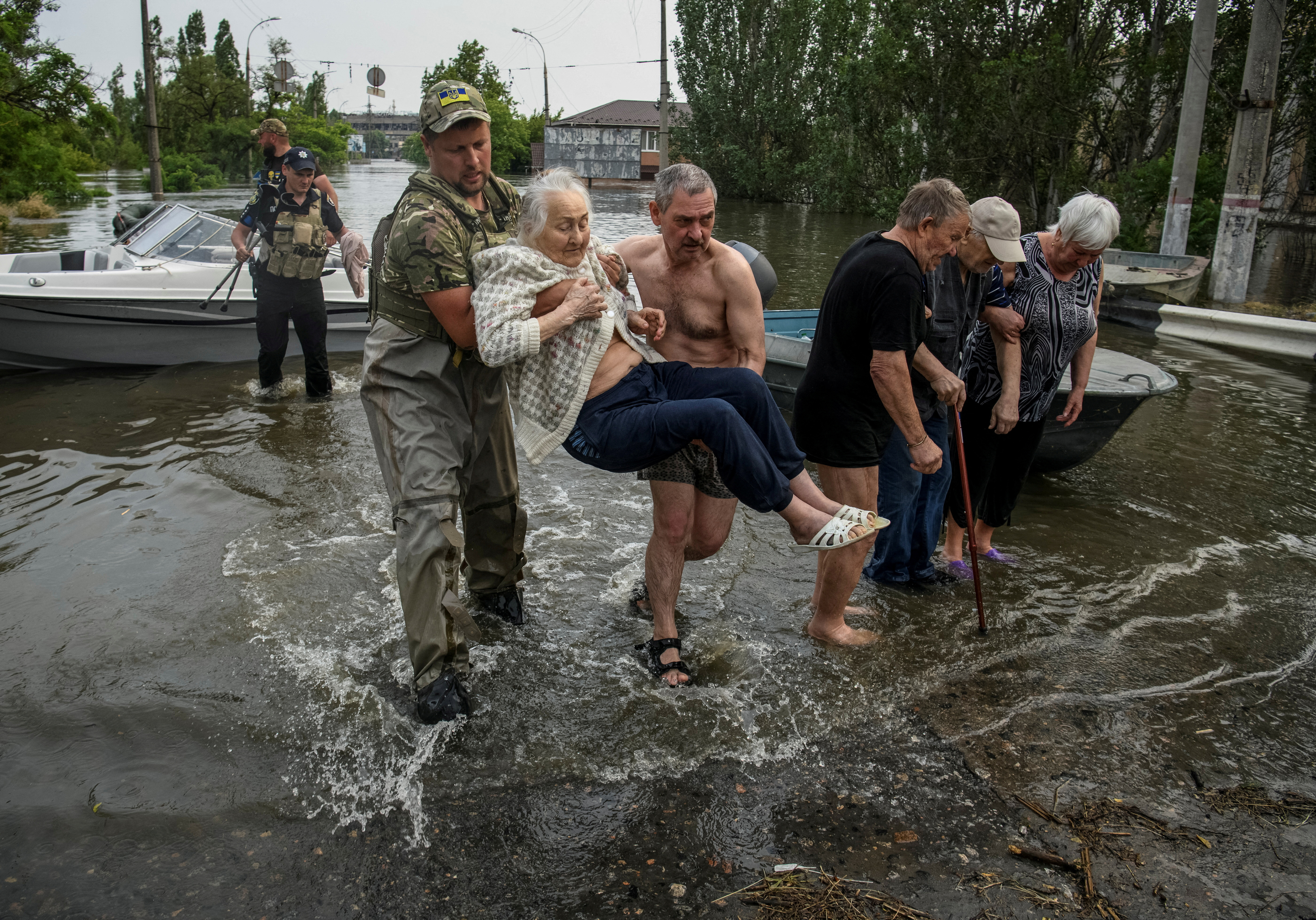 Two rescuers carrying a resident through flooding caused by the breach of the Nova Kakhovka dam. The resident is elderly. The water is not deep but is moving fast. Other people are also being rescued