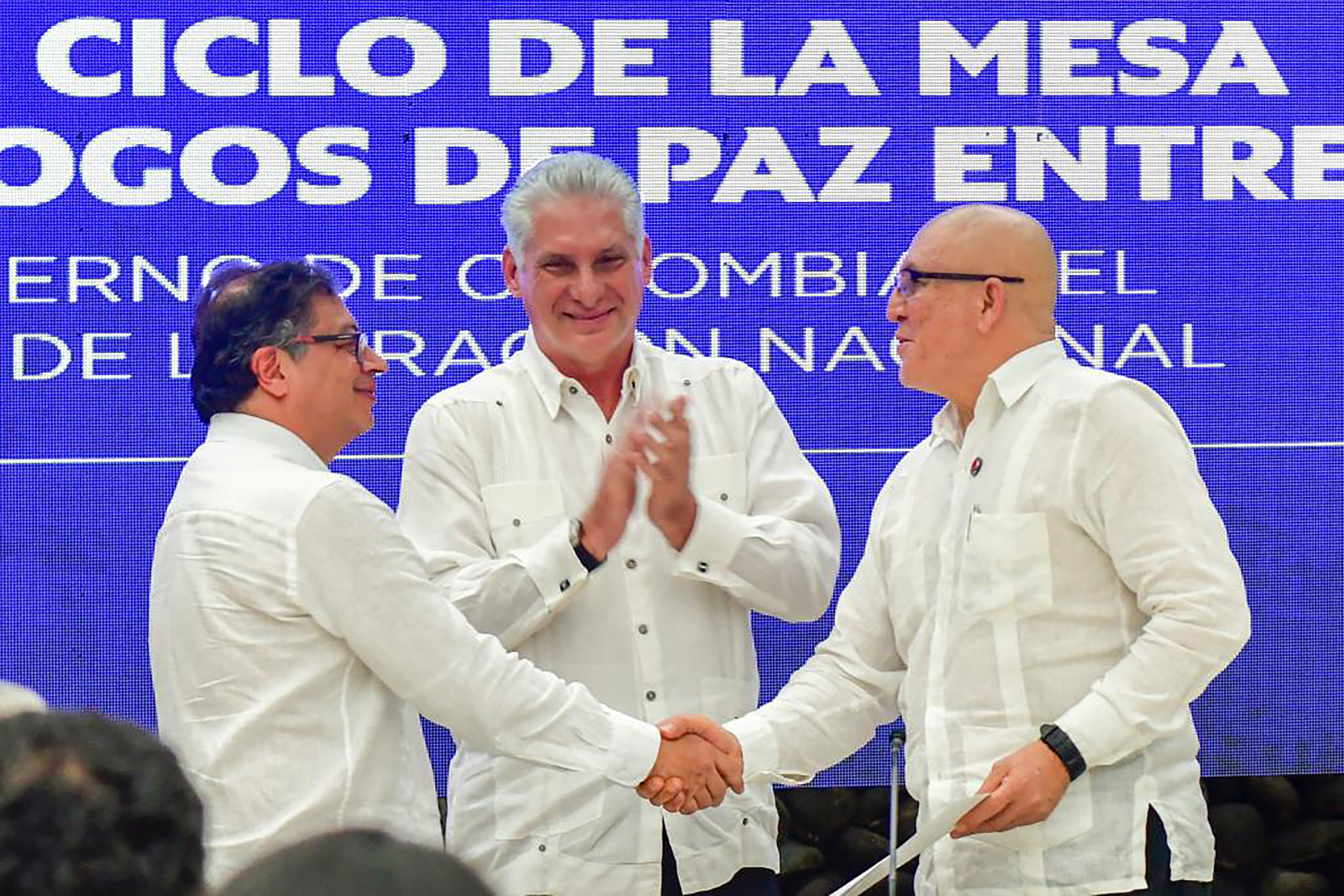 Two men, both dressed in white, shake hands, while another man claps behind them.